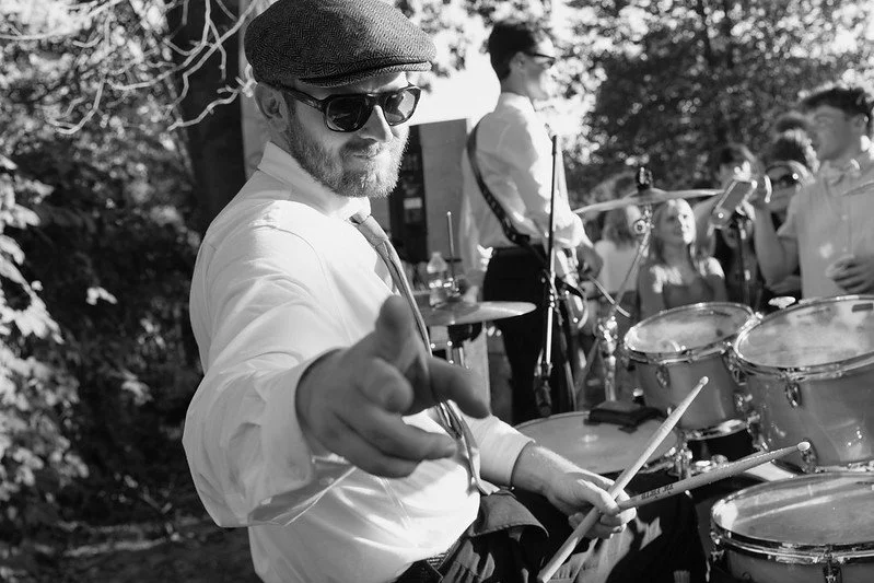 Man with beard wearing sunglasses, a hat, and white shirt, playing drums outdoors during a live music event.