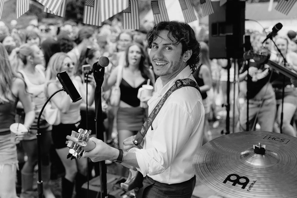 A musician with curly hair, wearing a white shirt, playing an electric guitar and smiling at the camera, surrounded by a crowd at a lively outdoor event with striped umbrellas.