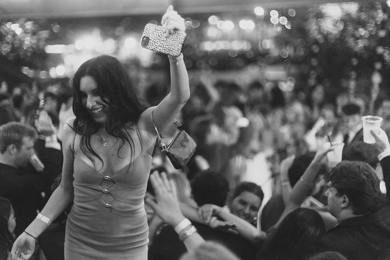 Woman dancing and smiling at party with crowd and drinks in hand, black and white photo.
