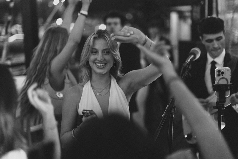 A young woman smiling and raising her hand at a social gathering or party, with people around her. She is holding a phone, and there is a microphone nearby, indicating a possible performance or karaoke setting.