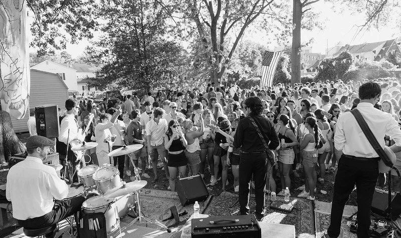 Musicians performing on stage with a drummer and a guitarist at an outdoor concert, surrounded by a large crowd of people in a park-like setting.