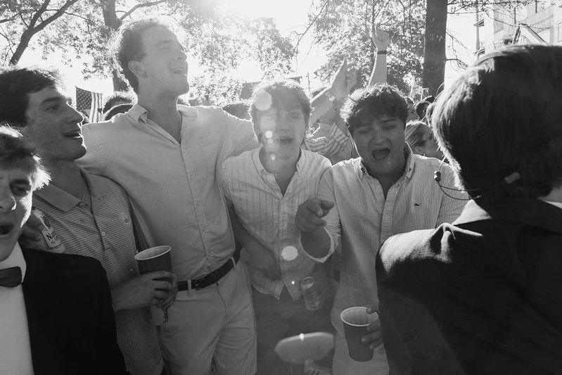 Group of young men smiling and celebrating outdoors, some holding cups, in a sunny park or street with trees and buildings in the background.