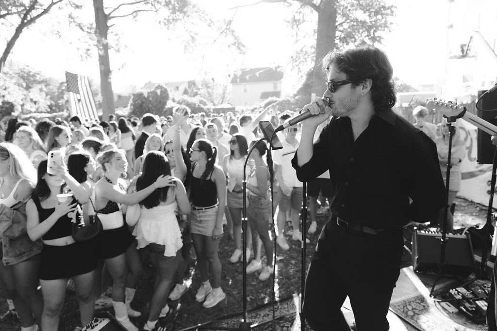 A man singing into a microphone on an outdoor stage with a crowd of people dancing and enjoying the music.
