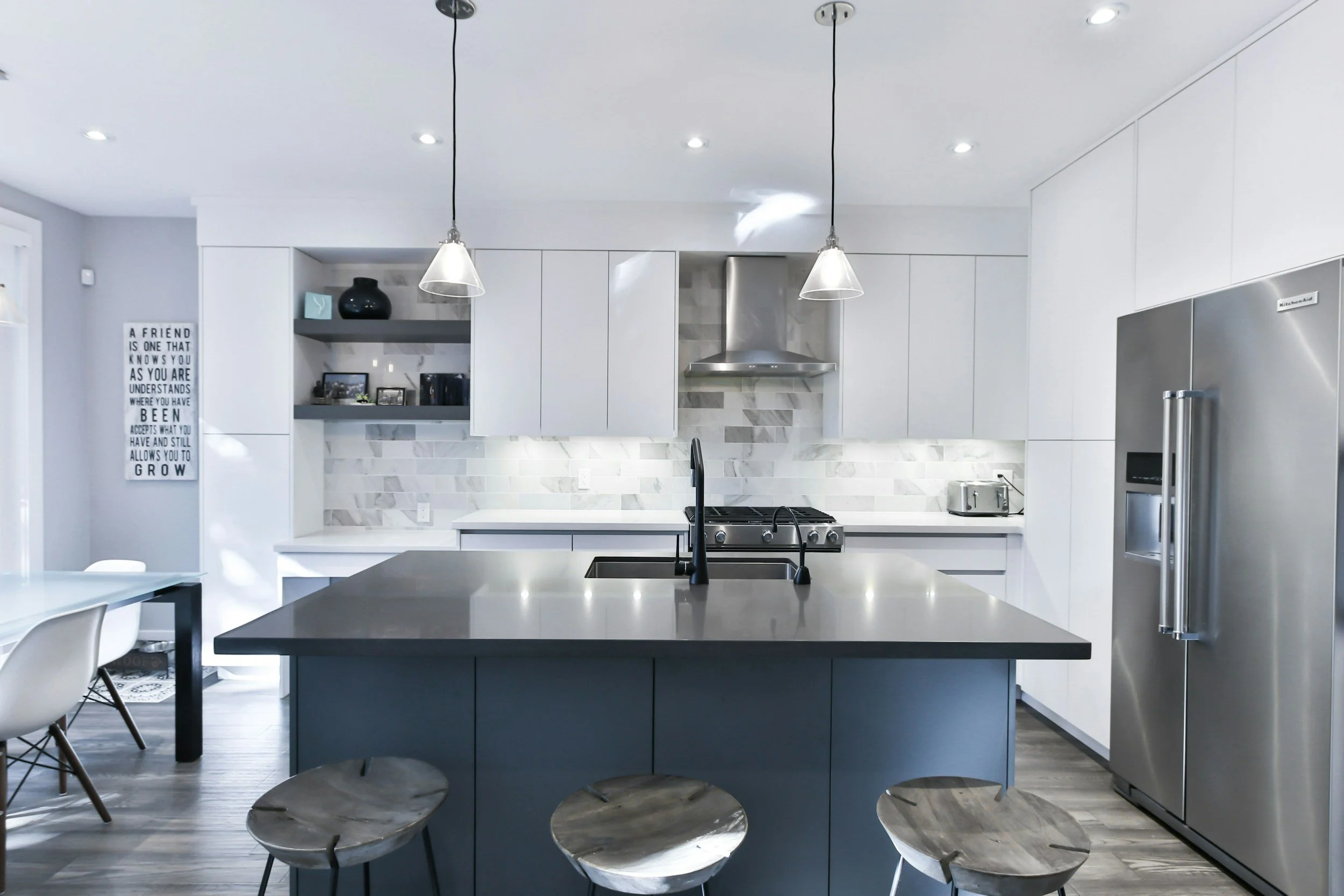 Modern kitchen with white cabinets, gray island, stainless steel refrigerator, and marble backsplash, illuminated by pendant and recessed lighting.