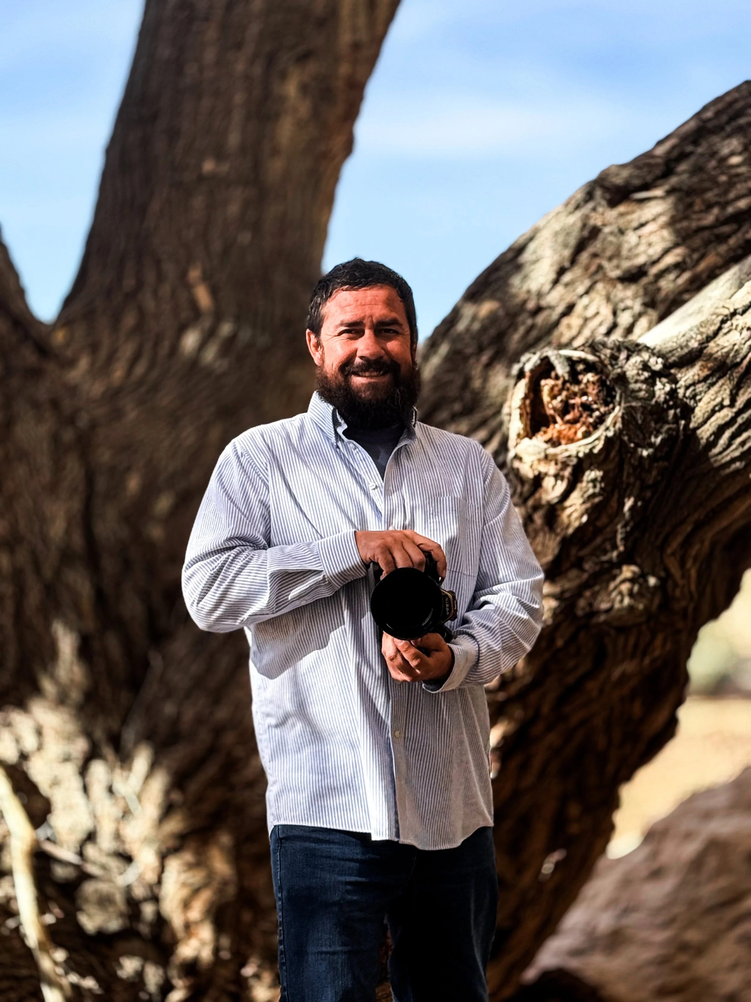 A man with dark hair and beard stands outdoors holding a camera, in front of a large tree trunk with branches and blue sky in the background.
