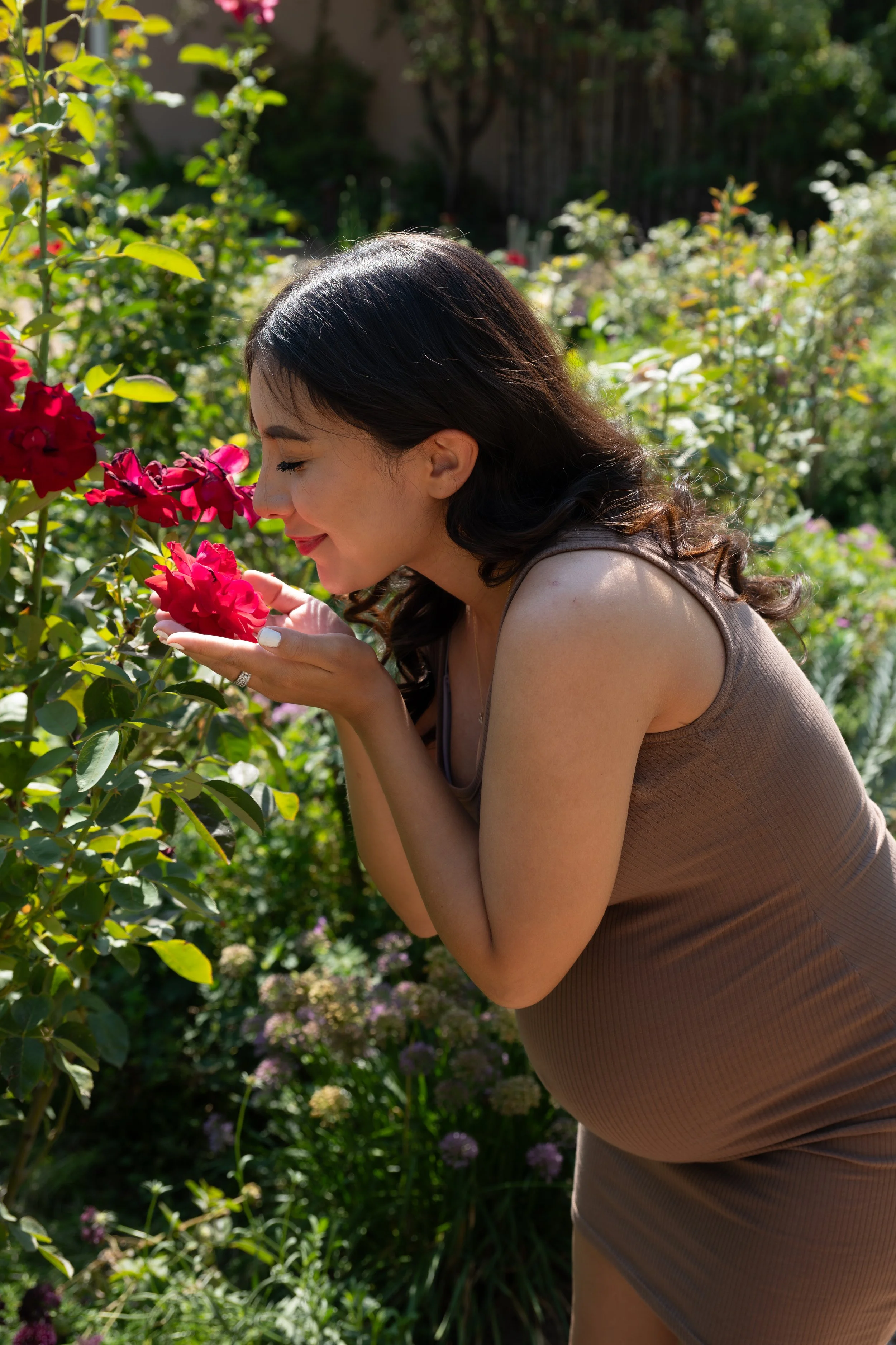 A woman with dark hair and a tank top is smelling pink roses in a garden.