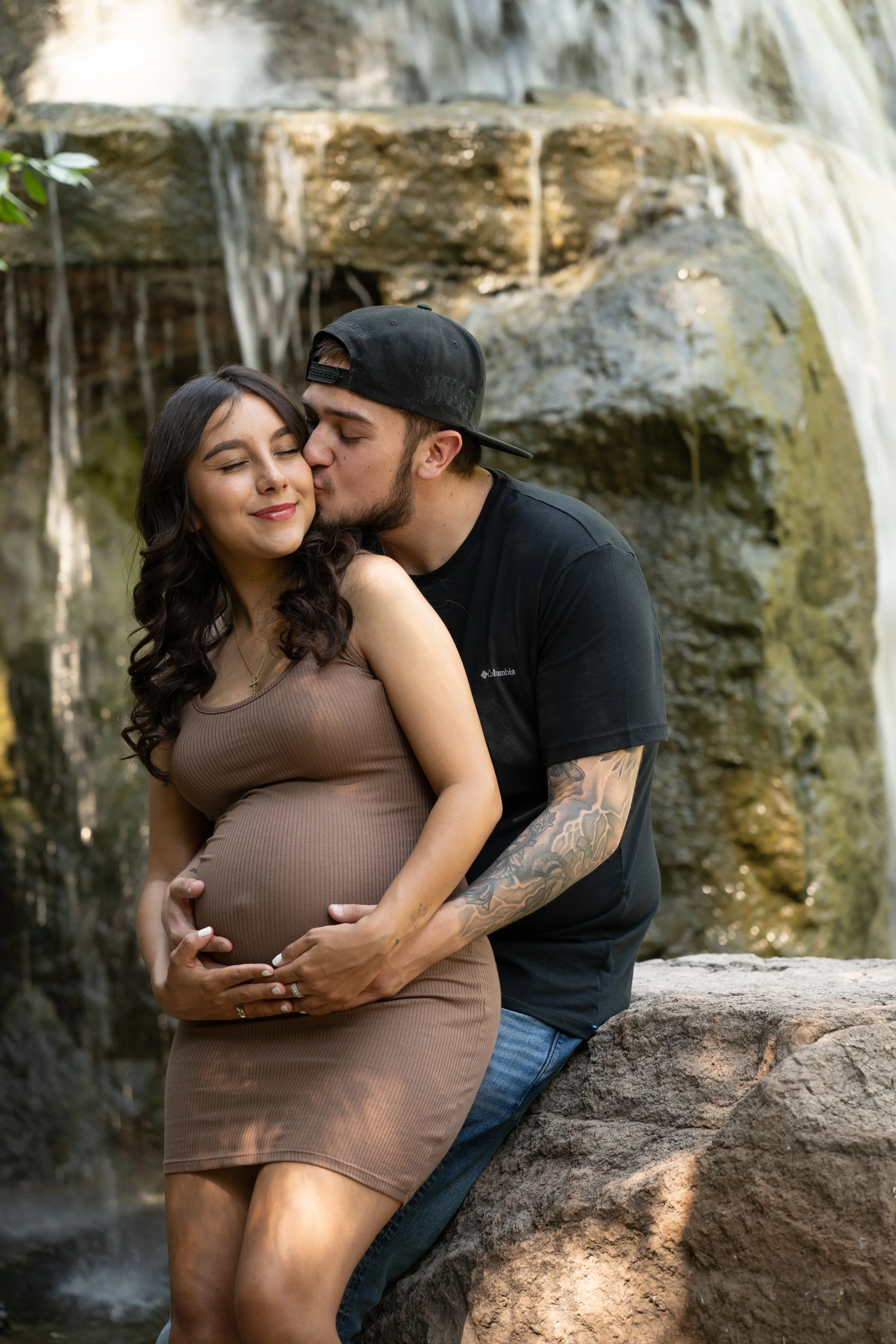 A pregnant woman in a beige dress is being kissed on the cheek by a man in a black T-shirt and baseball cap, sitting on a rock next to a waterfall.