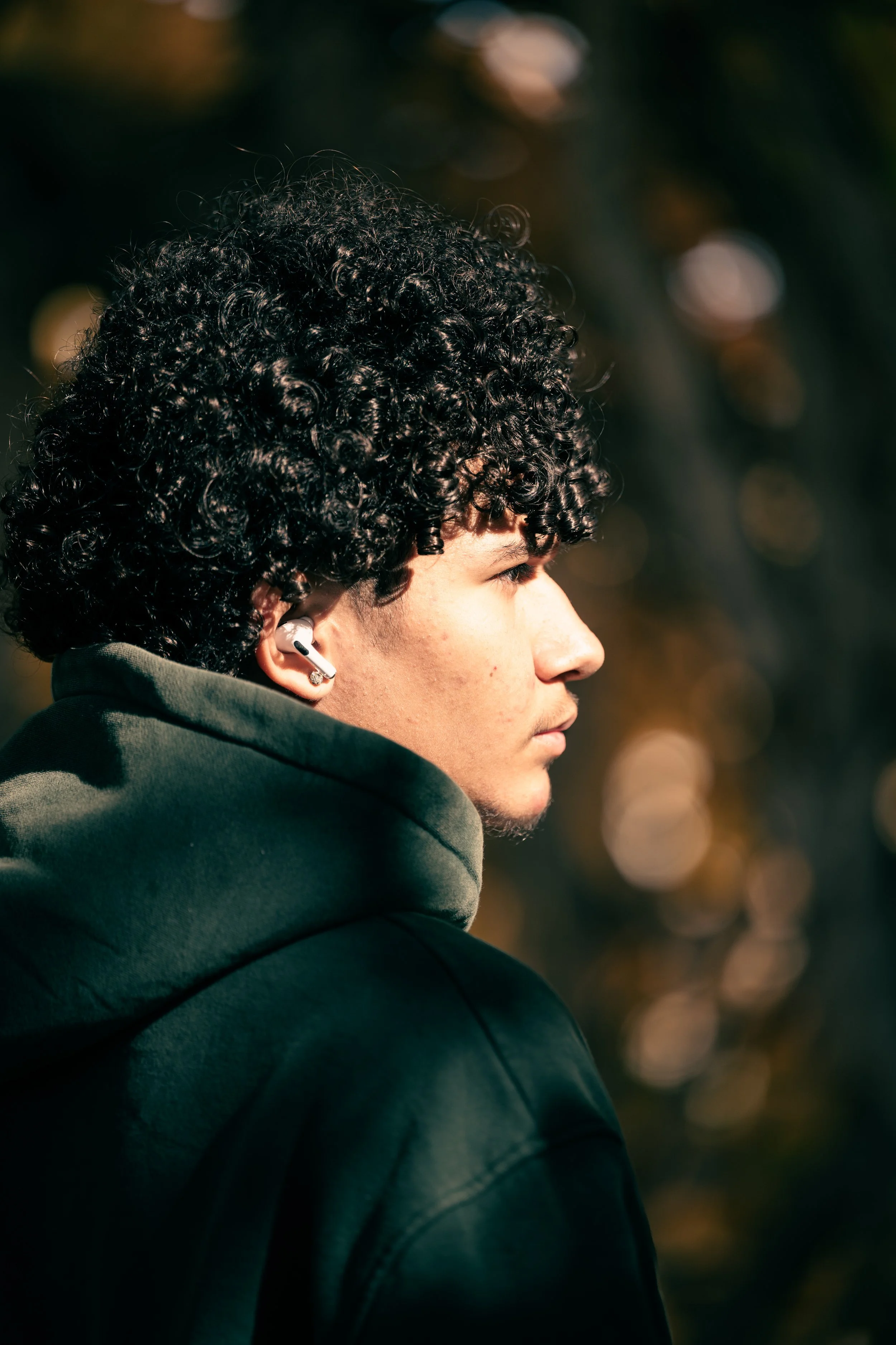A young man with curly black hair wearing a green hoodie and wireless earbuds, looking to the right, outdoors with a blurry background of trees and sunlight.