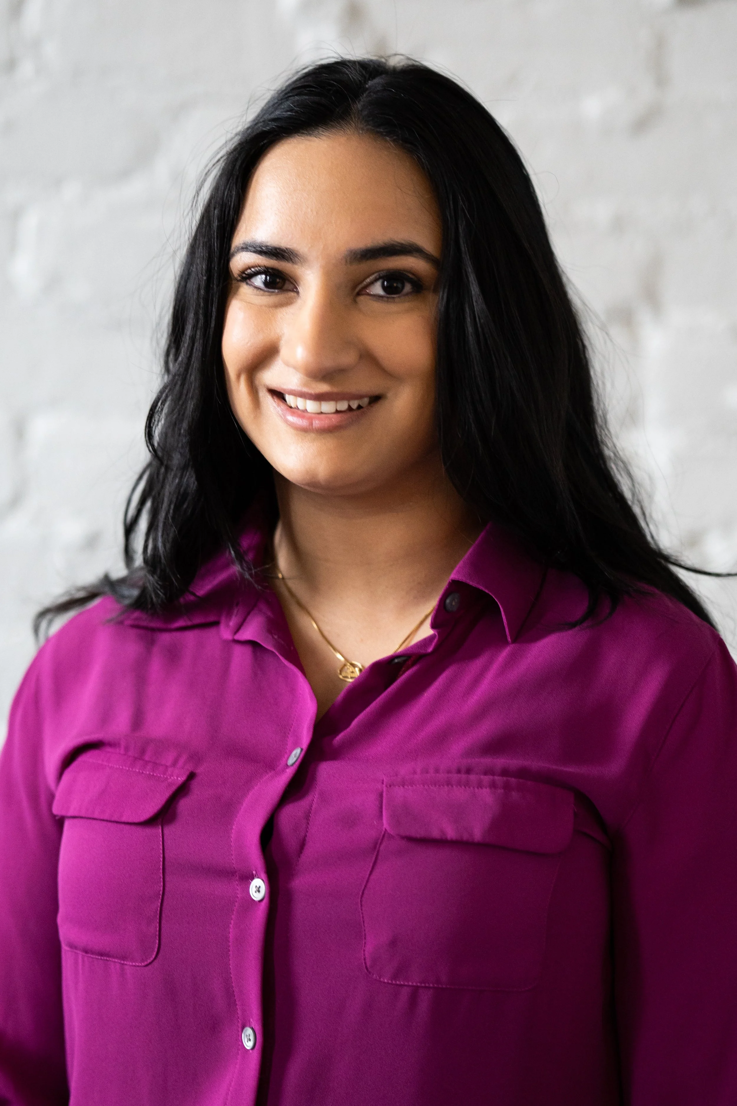 A woman with long black hair, wearing a purple shirt and gold necklace, smiling against a white brick wall background.