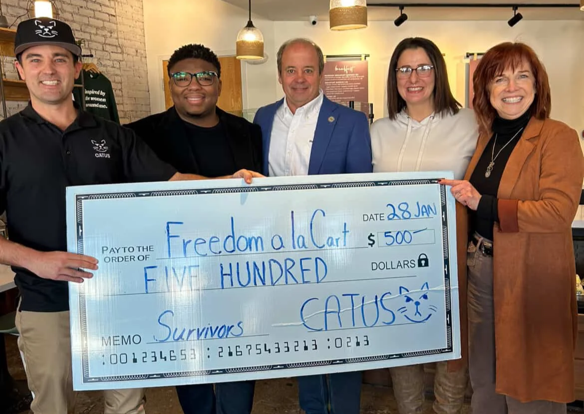 Group of five people holding a large ceremonial check for $500 made out to Freedom la Cart, dated January 28, 2023, with the memo 'Survivors,' in a cafe or restaurant setting.