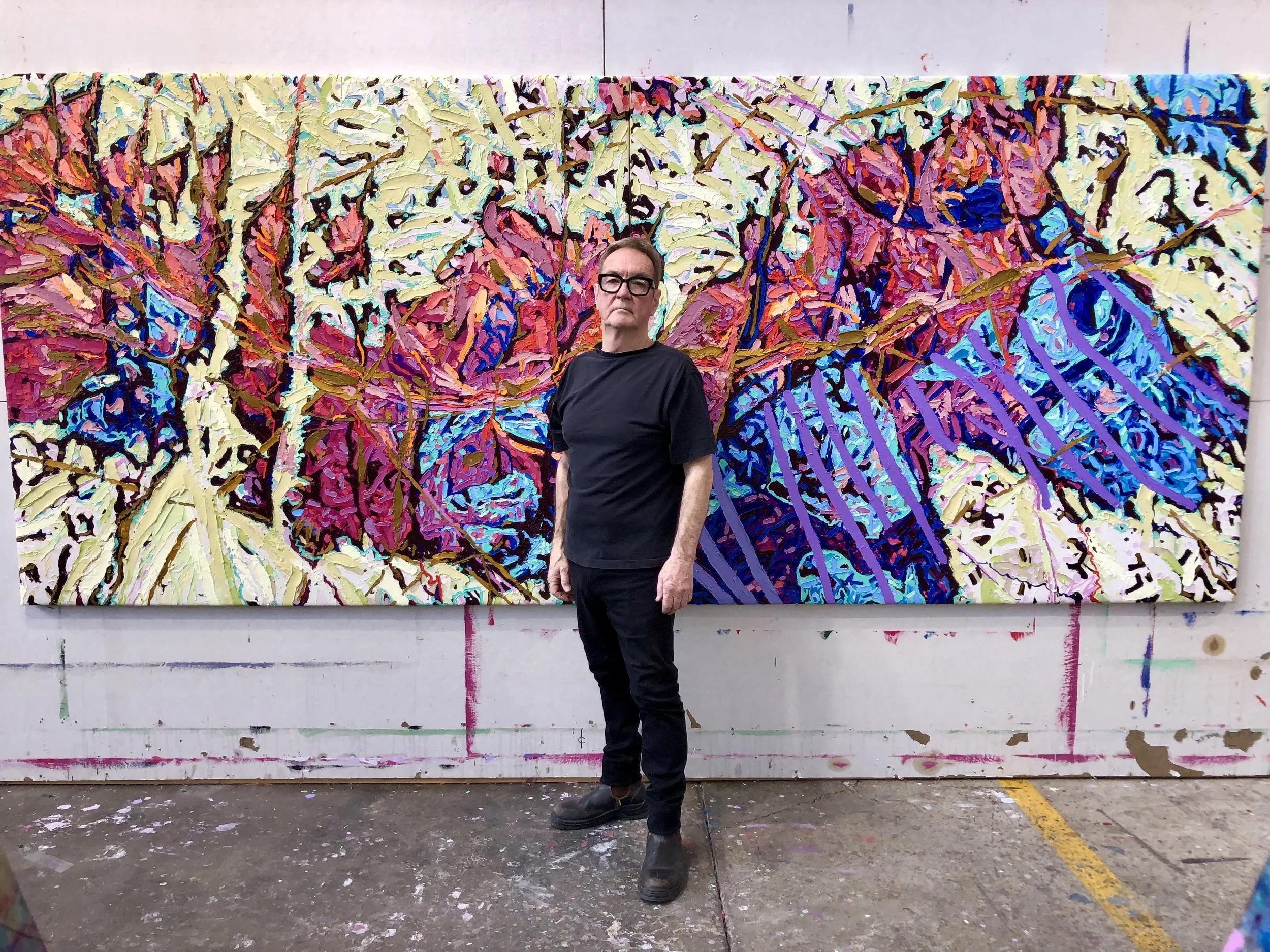 A man in black clothing and glasses standing in front of a large colorful abstract painting. The painting features bold, textured strokes in pink, blue, yellow, purple, and black colors.