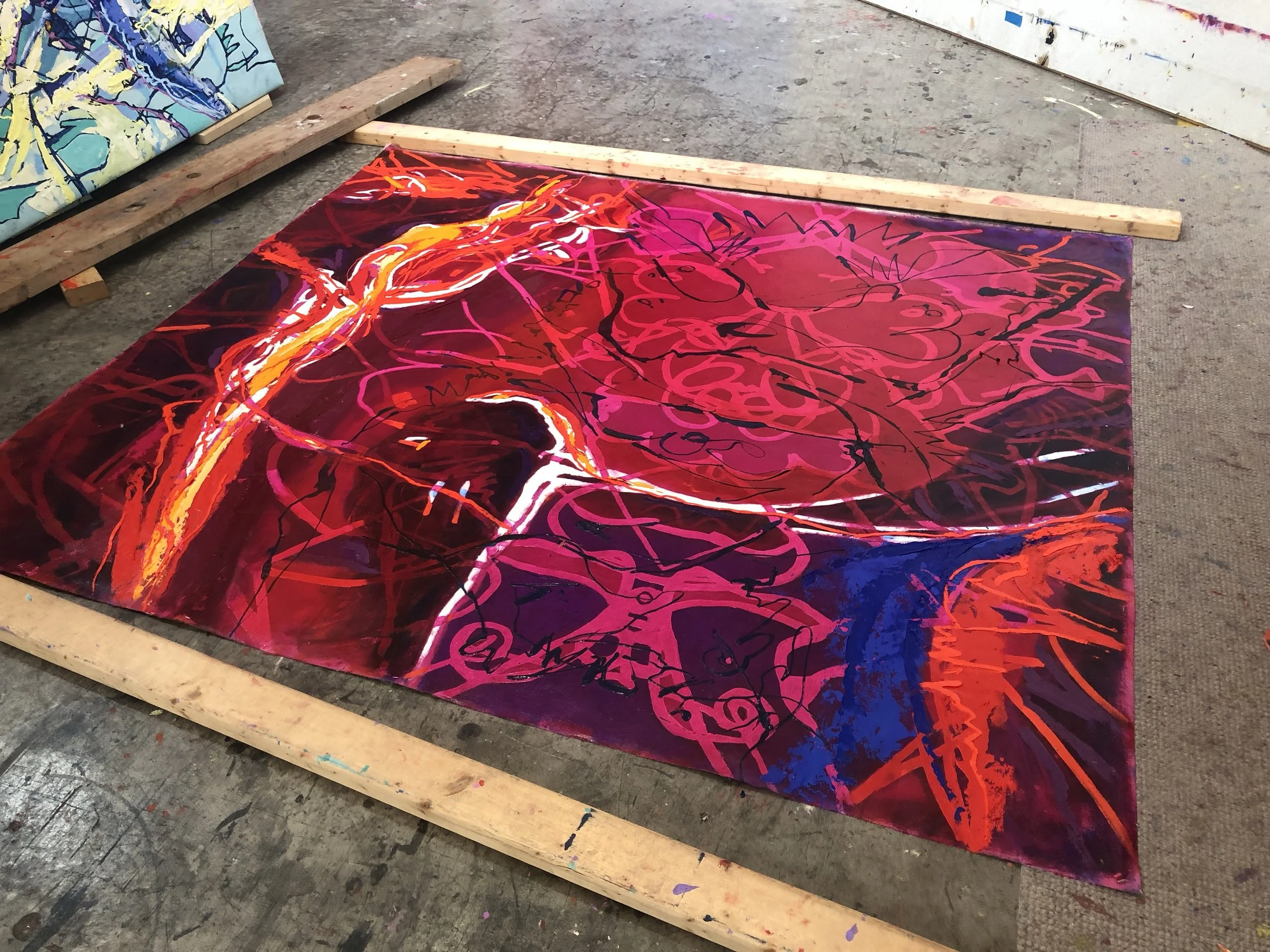 A colorful abstract painting with vibrant reds, pinks, blues, and oranges, featuring swirling and layered lines and shapes, laying on the floor of an art studio.