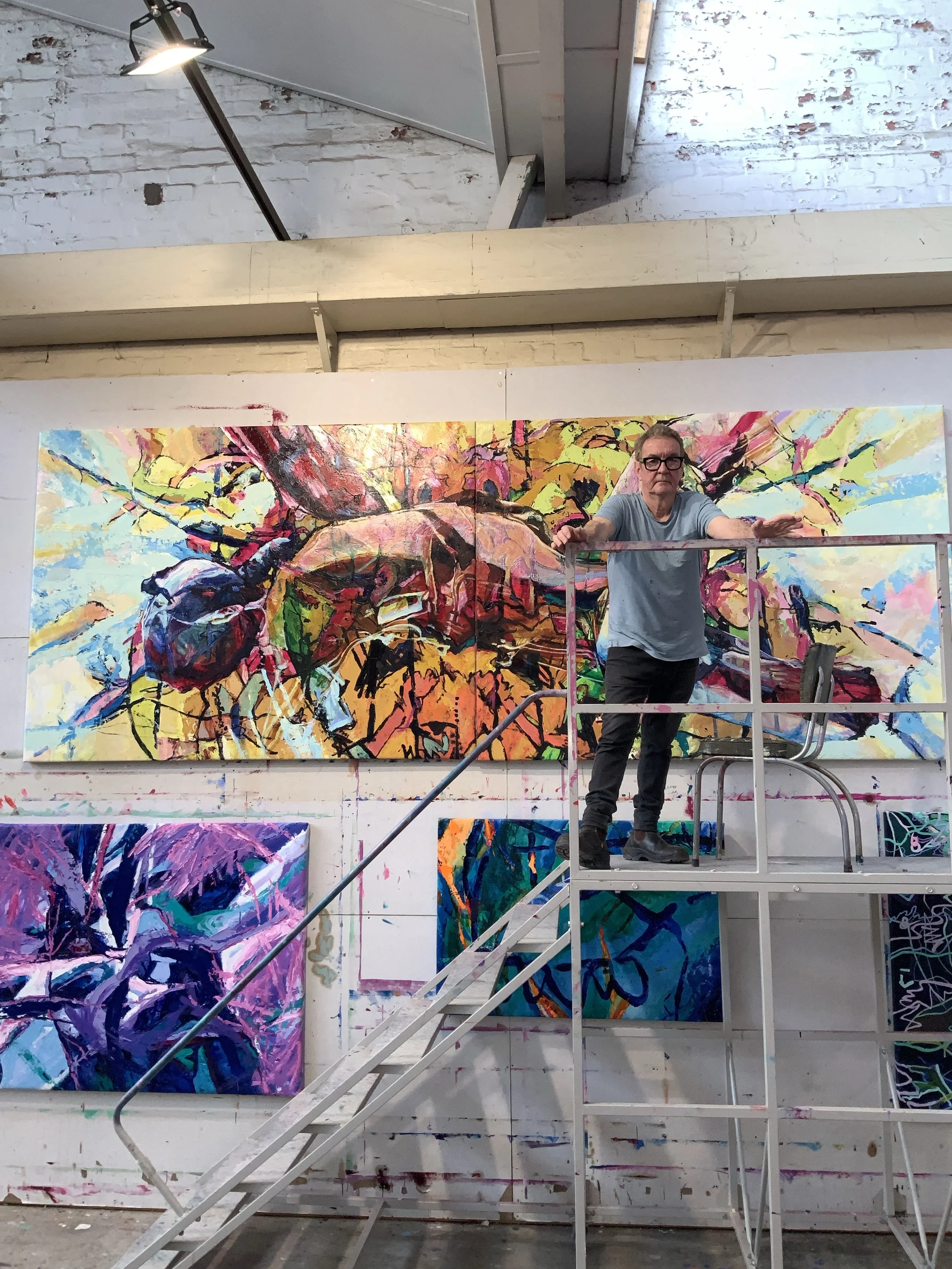 An artist in a blue t-shirt and glasses stands on a metal scaffold in front of a large, colorful abstract painting on a white wall in an art studio.