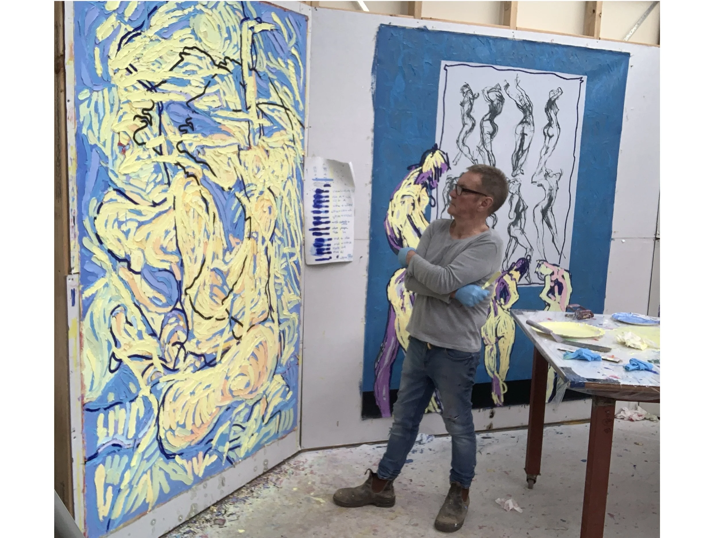 An artist with glasses and gray hair, wearing a gray long-sleeve shirt, blue gloves, jeans, and brown shoes, stands in an art studio with arms crossed, looking at large abstract paintings on the walls. The studio has a table with art supplies and a p