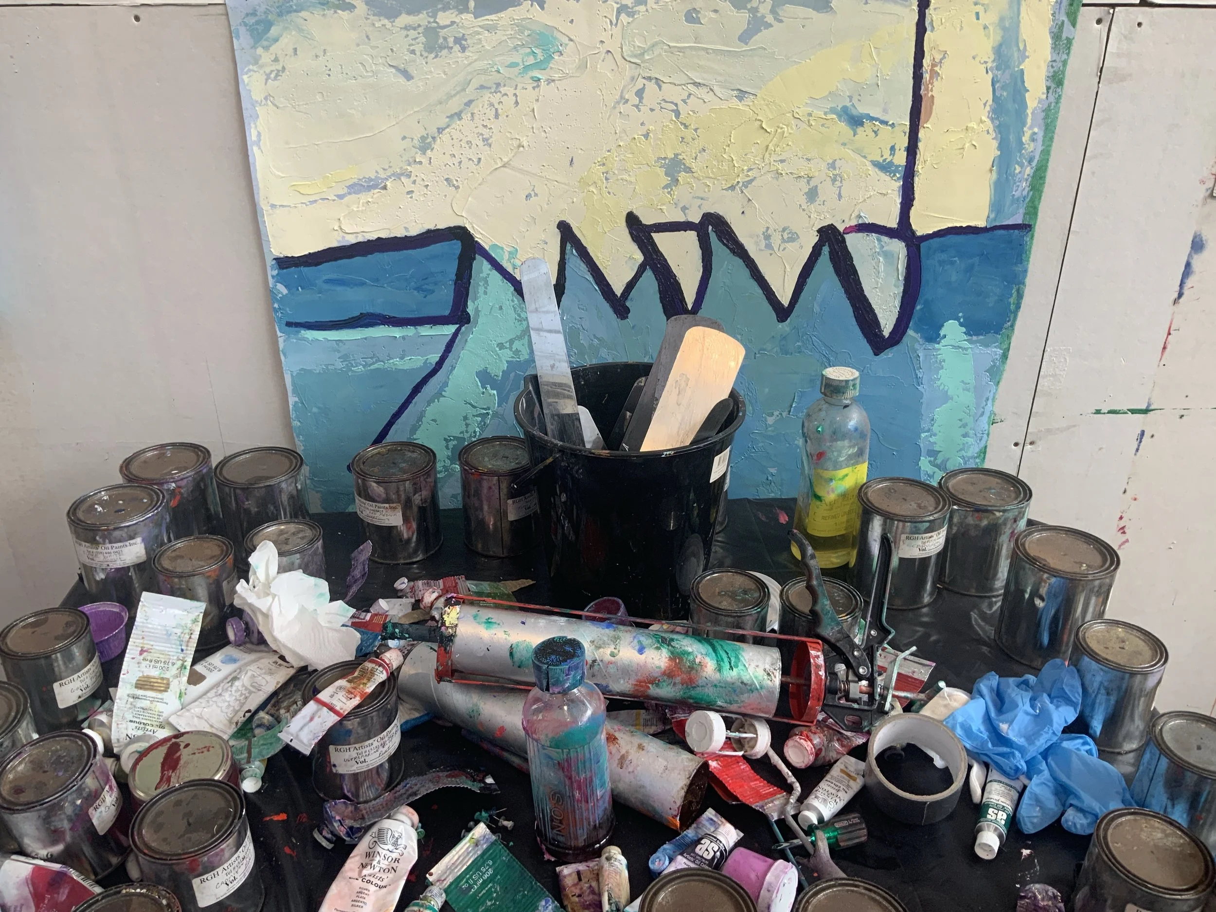 A cluttered artist's workspace with paint cans, brushes, and used supplies, in front of an abstract painting with blue, yellow, and purple colors.