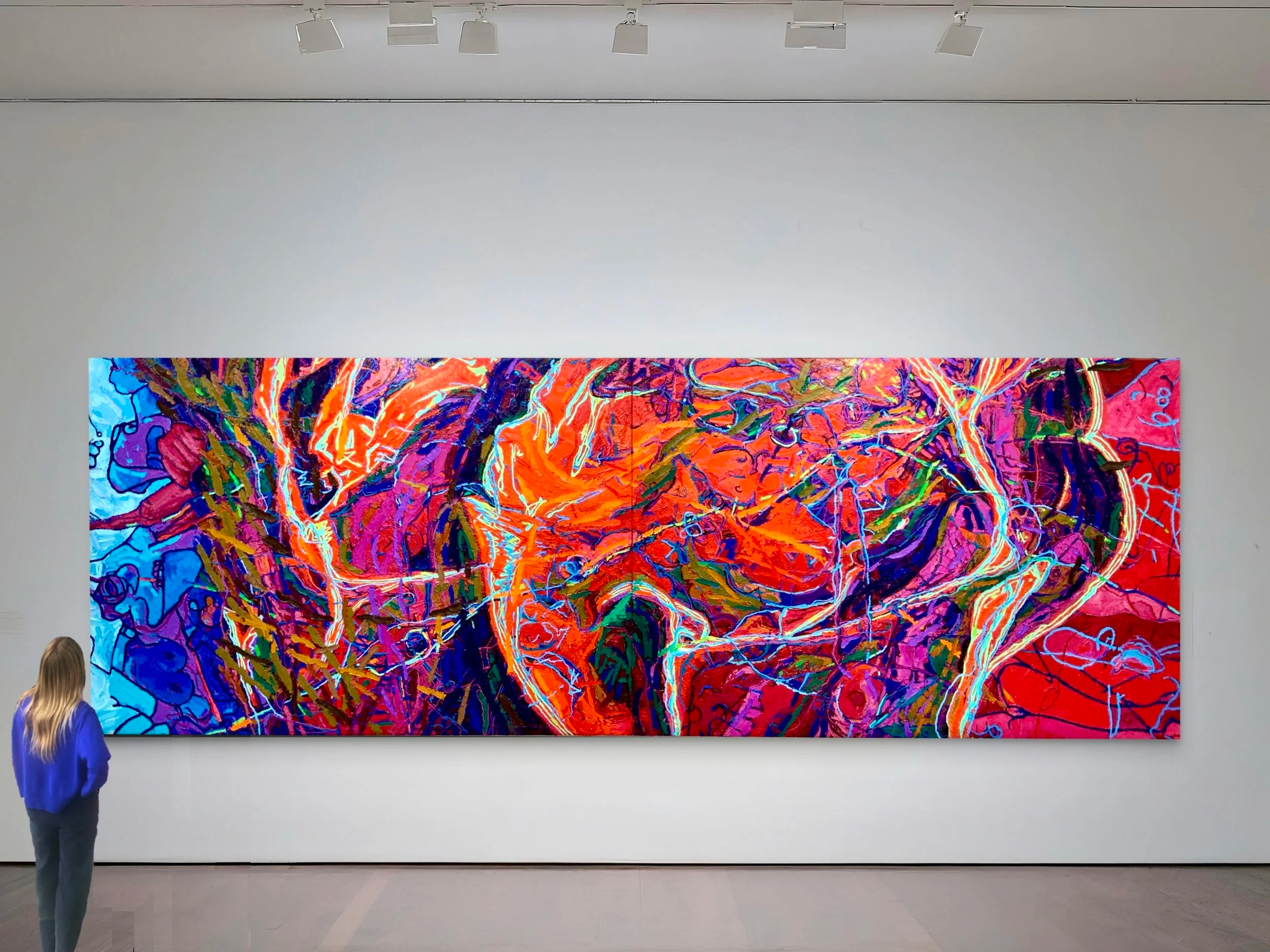 An abstract colorful painting exhibited in a gallery on a white wall, with a woman observing it.