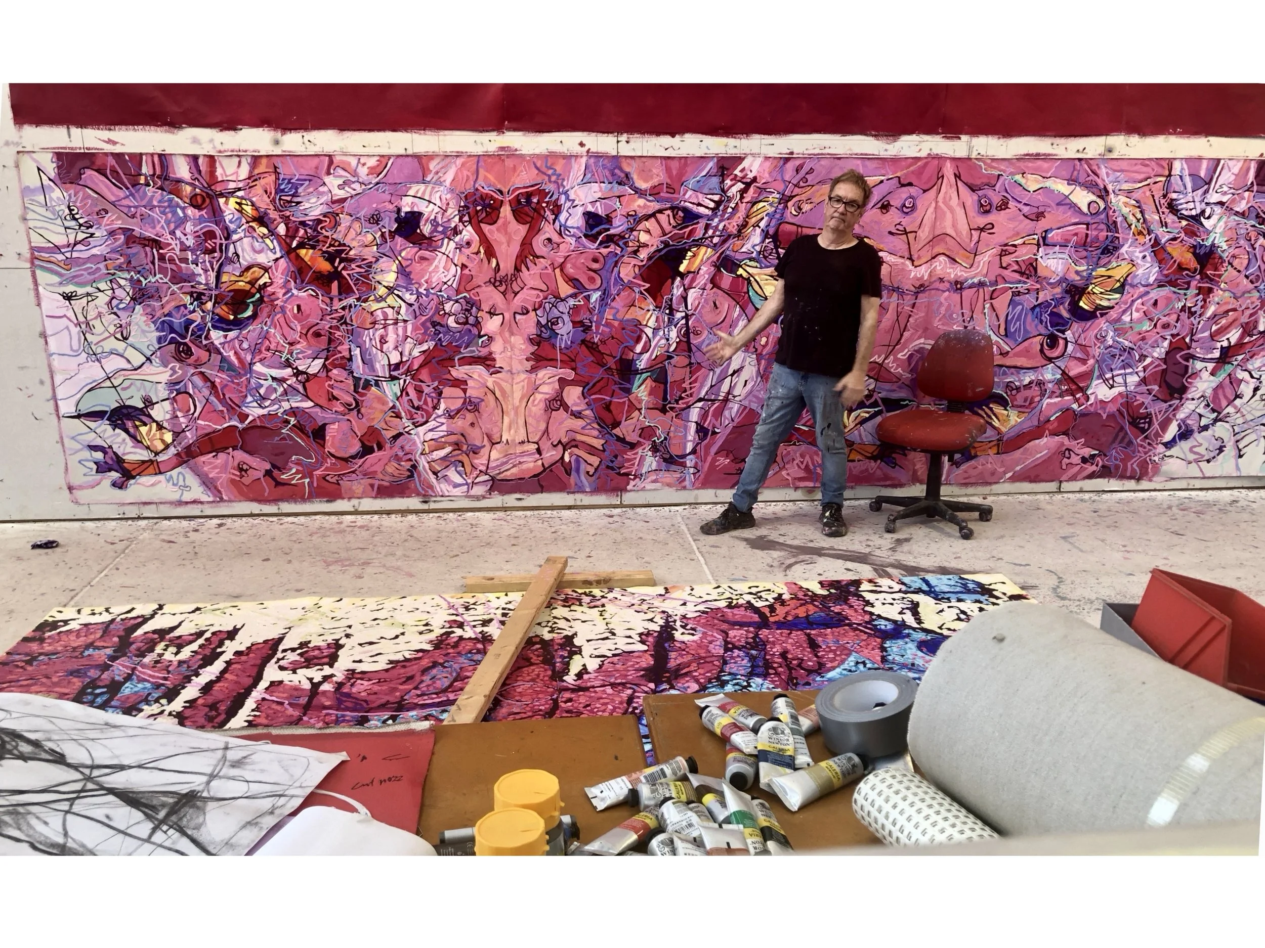 An artist standing in an art studio in front of a large pink abstract mural with intricate lines and vibrant colors. The studio has art supplies, paint tubes, and canvases on the floor.