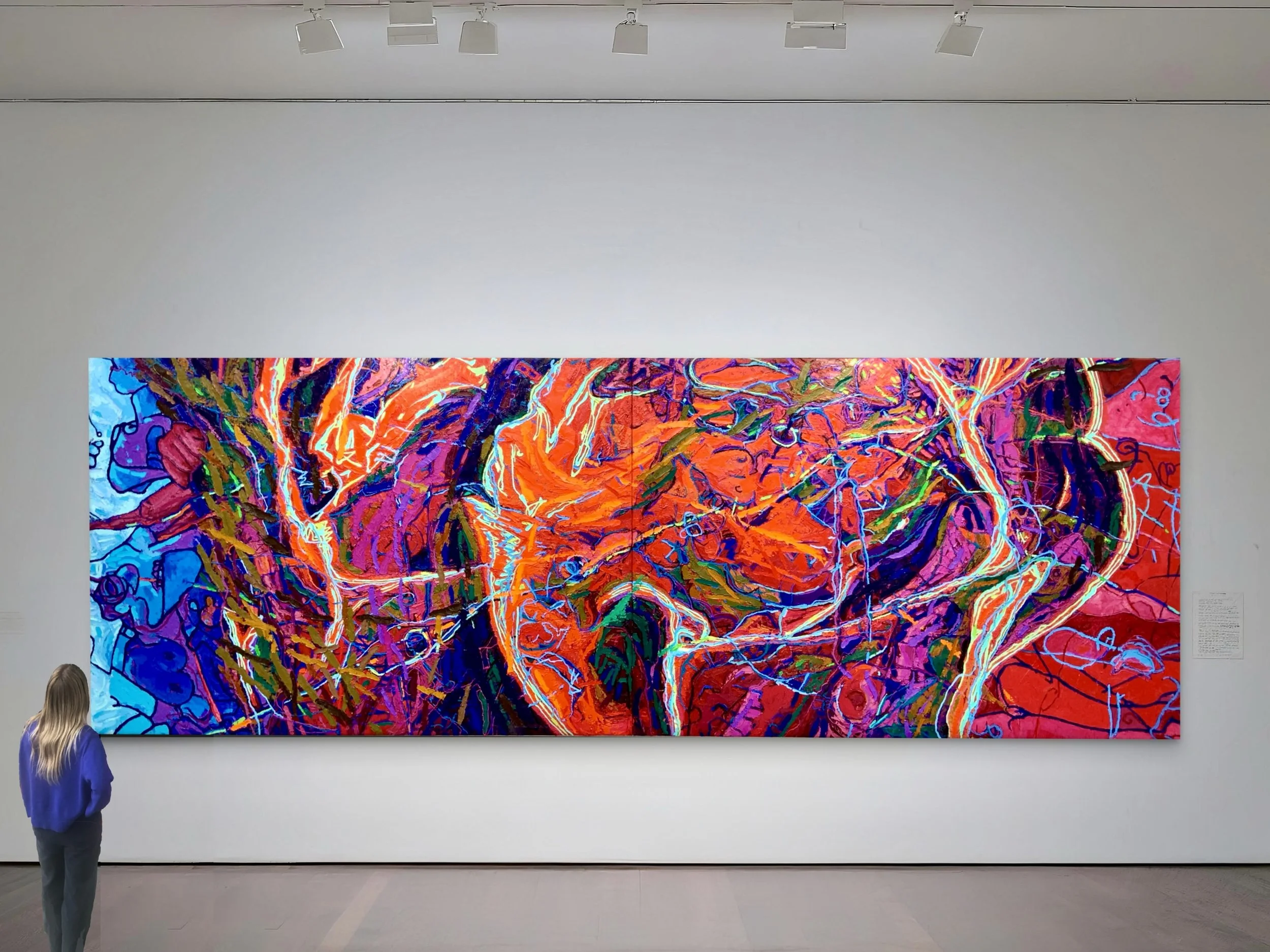 A large, colorful abstract painting displayed in a gallery. The artwork features vibrant, energetic strokes and swirling lines in reds, blues, oranges, and purples. A person with long blond hair, wearing a blue jacket, stands in front of the painting, observing it.