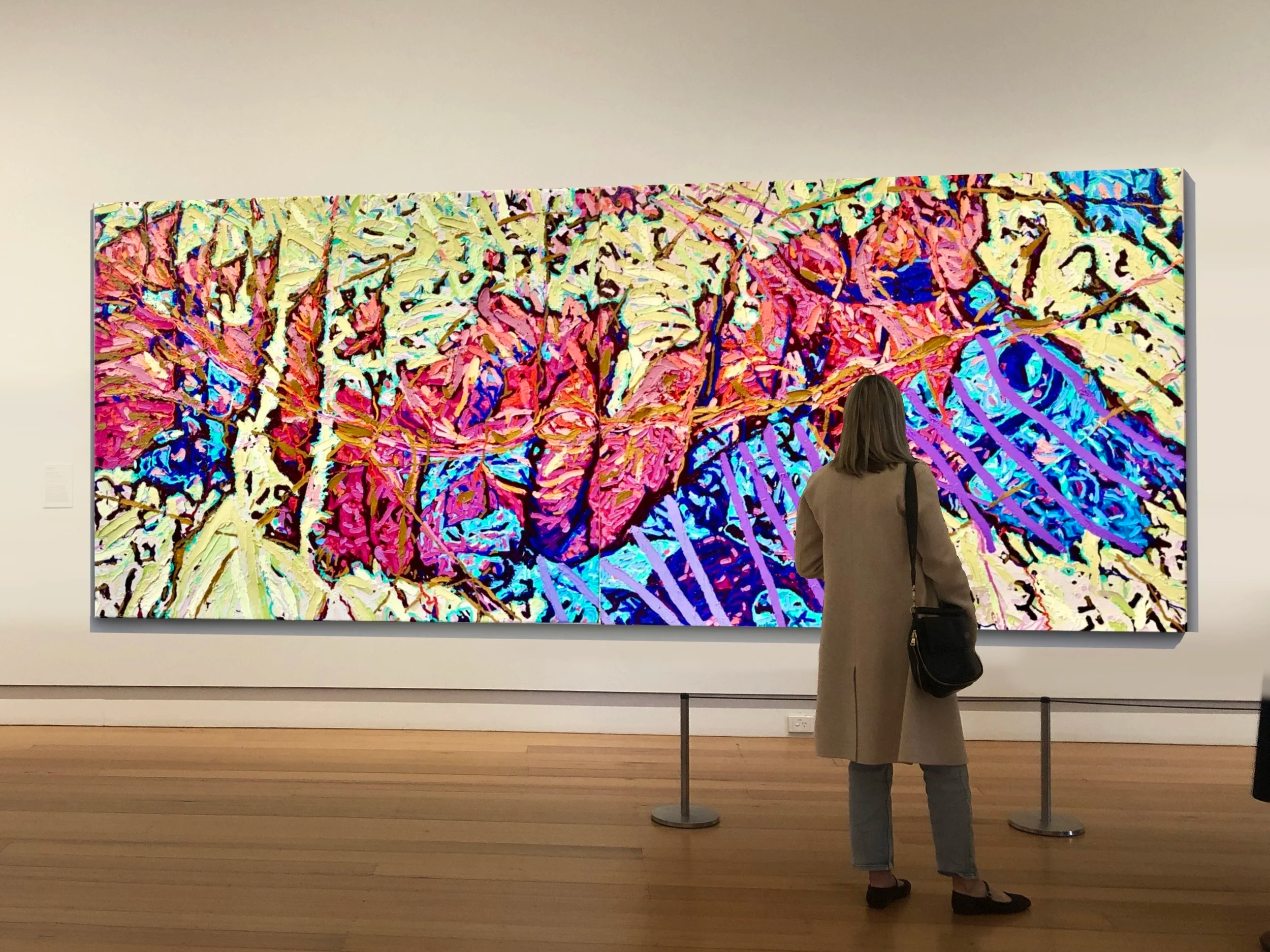 A woman in a beige coat and jeans stands in front of a colorful abstract painting in an art gallery, viewed from behind.