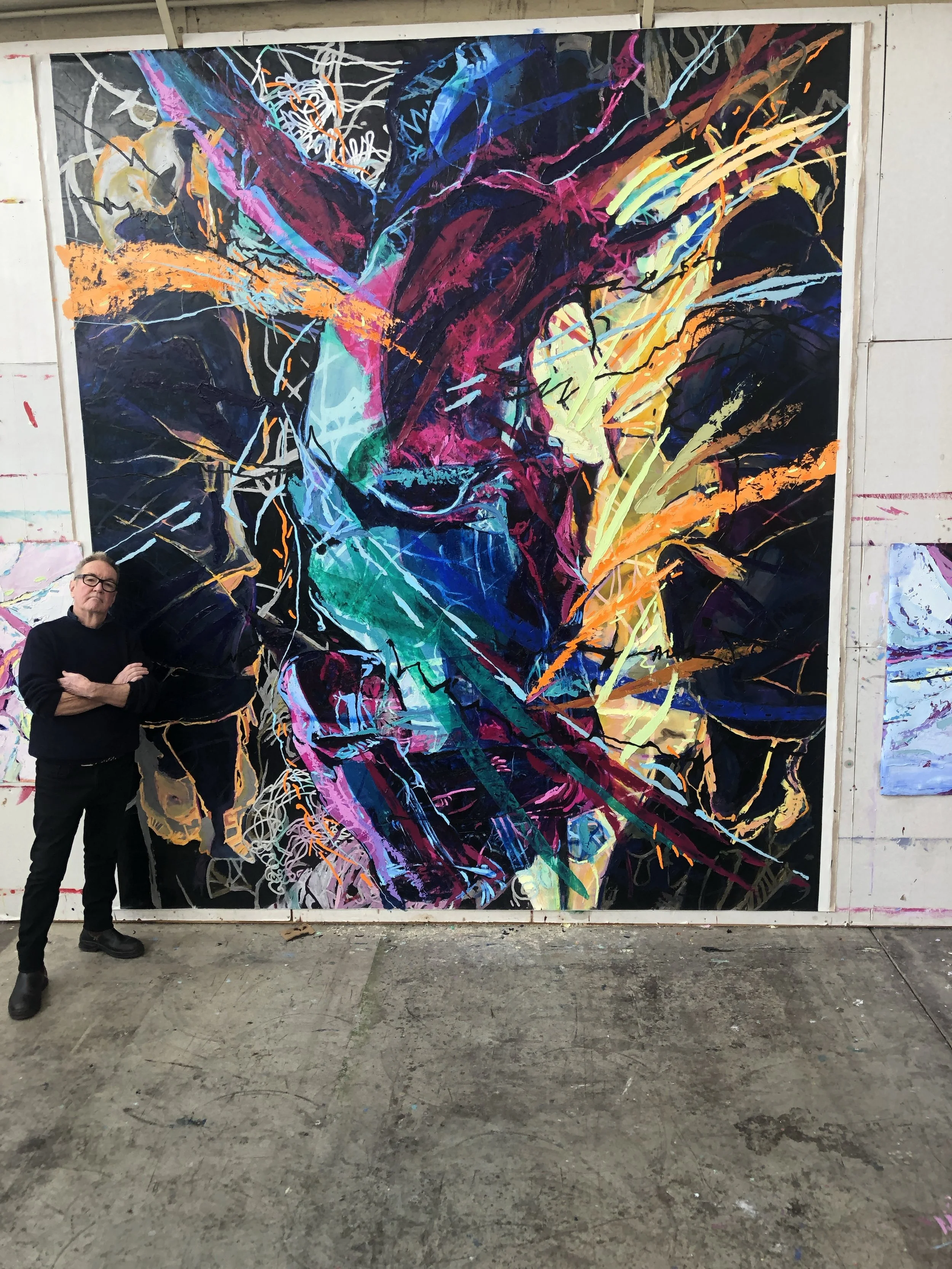 A man with glasses wearing a black shirt stands with his arms crossed next to a large vibrant abstract portrait of a dog's face painted with bright colors and dynamic brushstrokes, displayed on a wall.