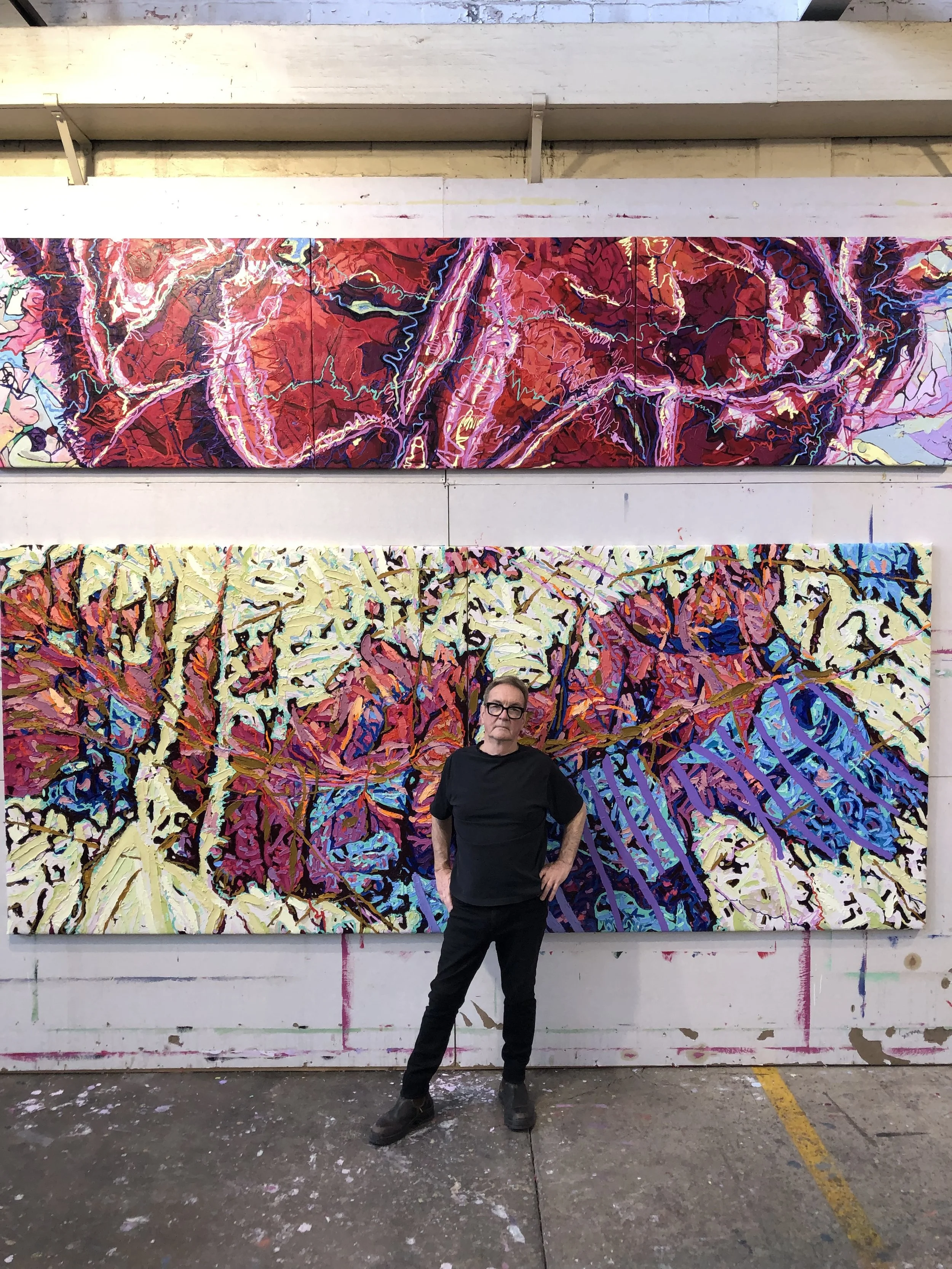An older man wearing black clothes and glasses standing in front of large colorful abstract paintings in an art studio.