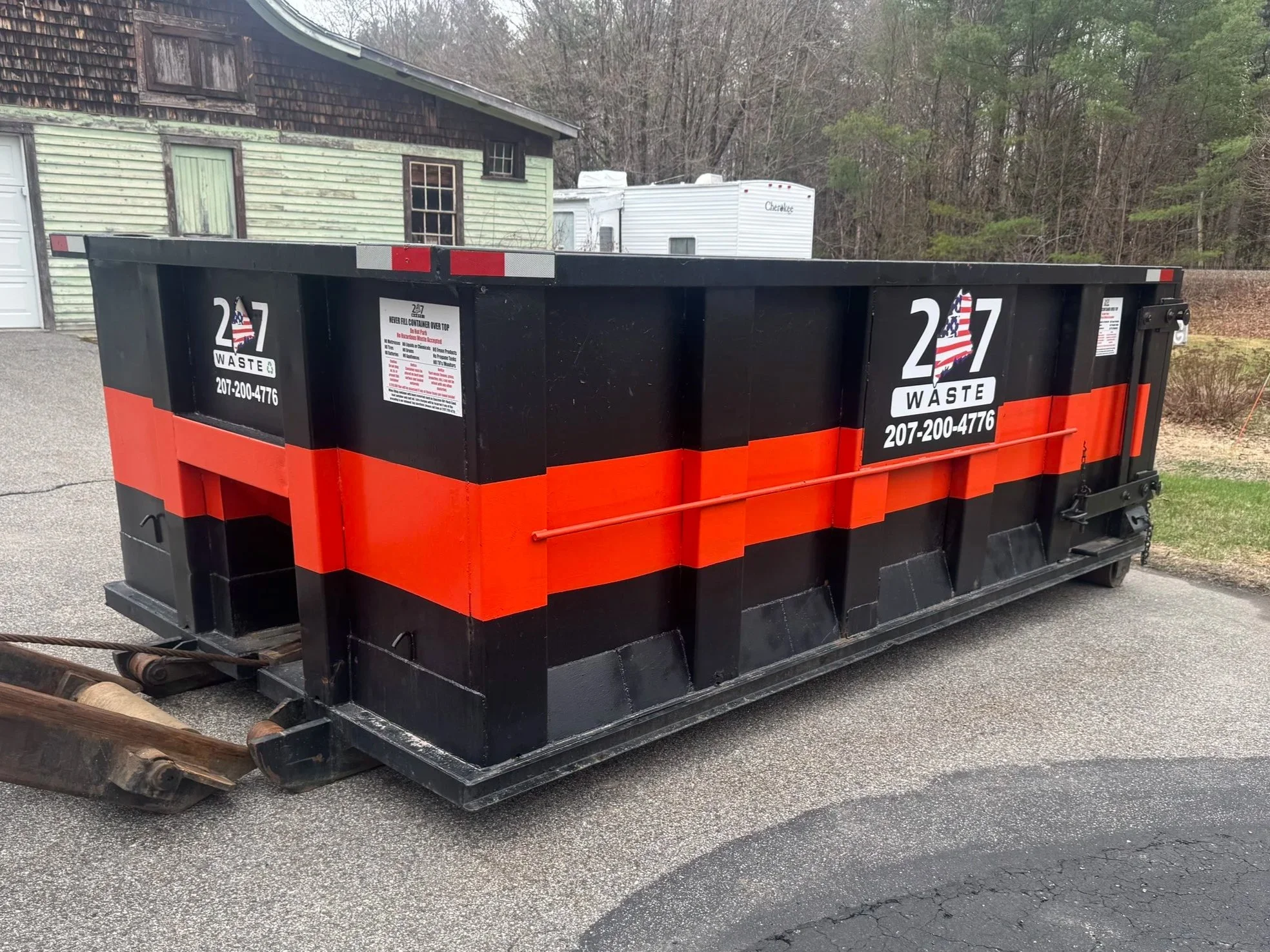 A two-story white house with an attached garage, surrounded by green lawn and trees, with a large orange dumpster on the driveway filled with trash.  207 Waste Poland Maine, Wast Management Maine