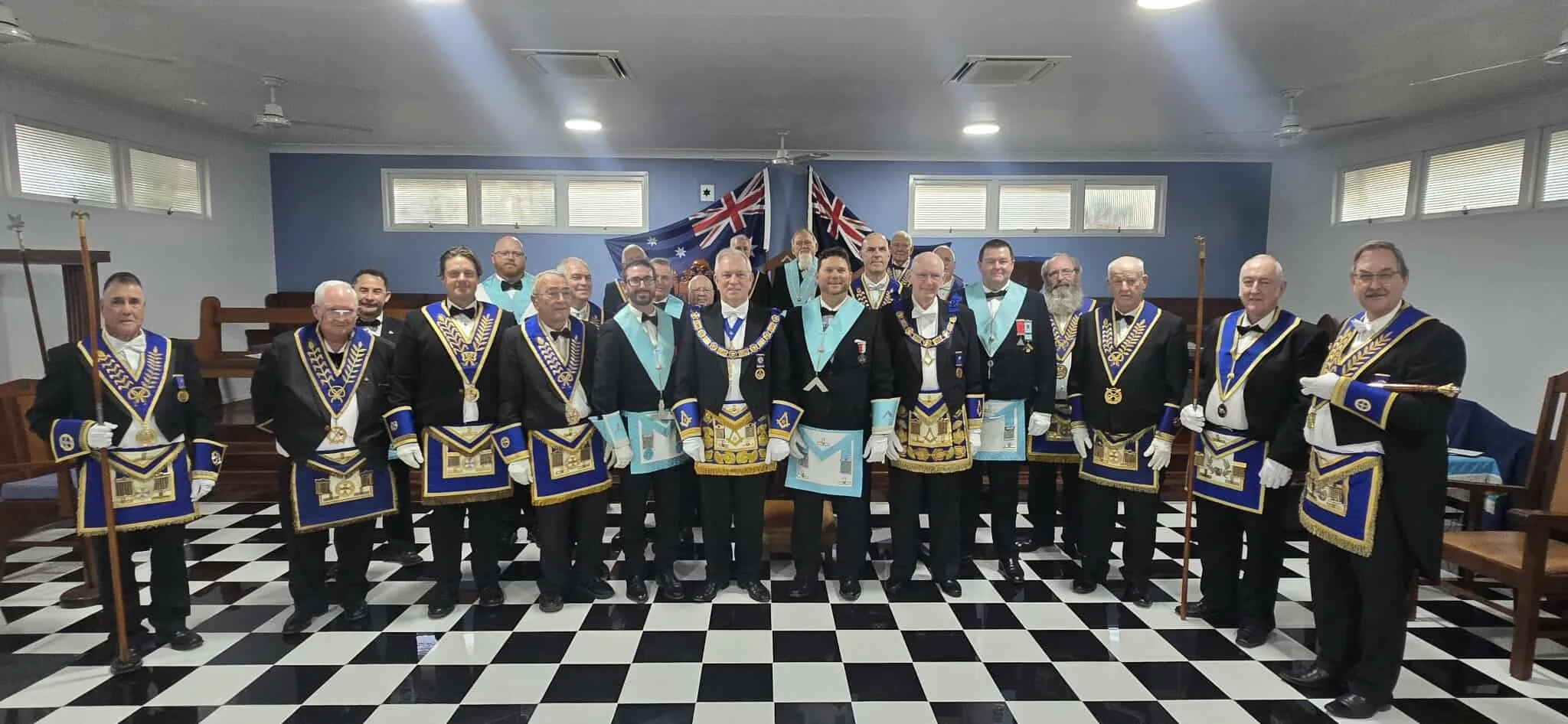 Group of men in formal Masonic regalia standing in a lodge room with Australian flags in the background.