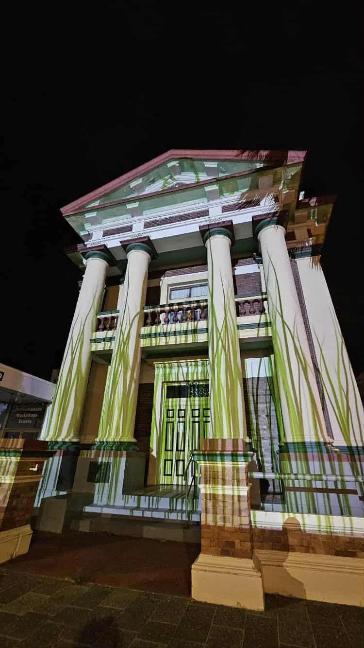 A large Masonic Hall in Mackay, Queensland illuminated at night with green and purple light projections, featuring tall columns, a balcony, and steps leading up to the front door.