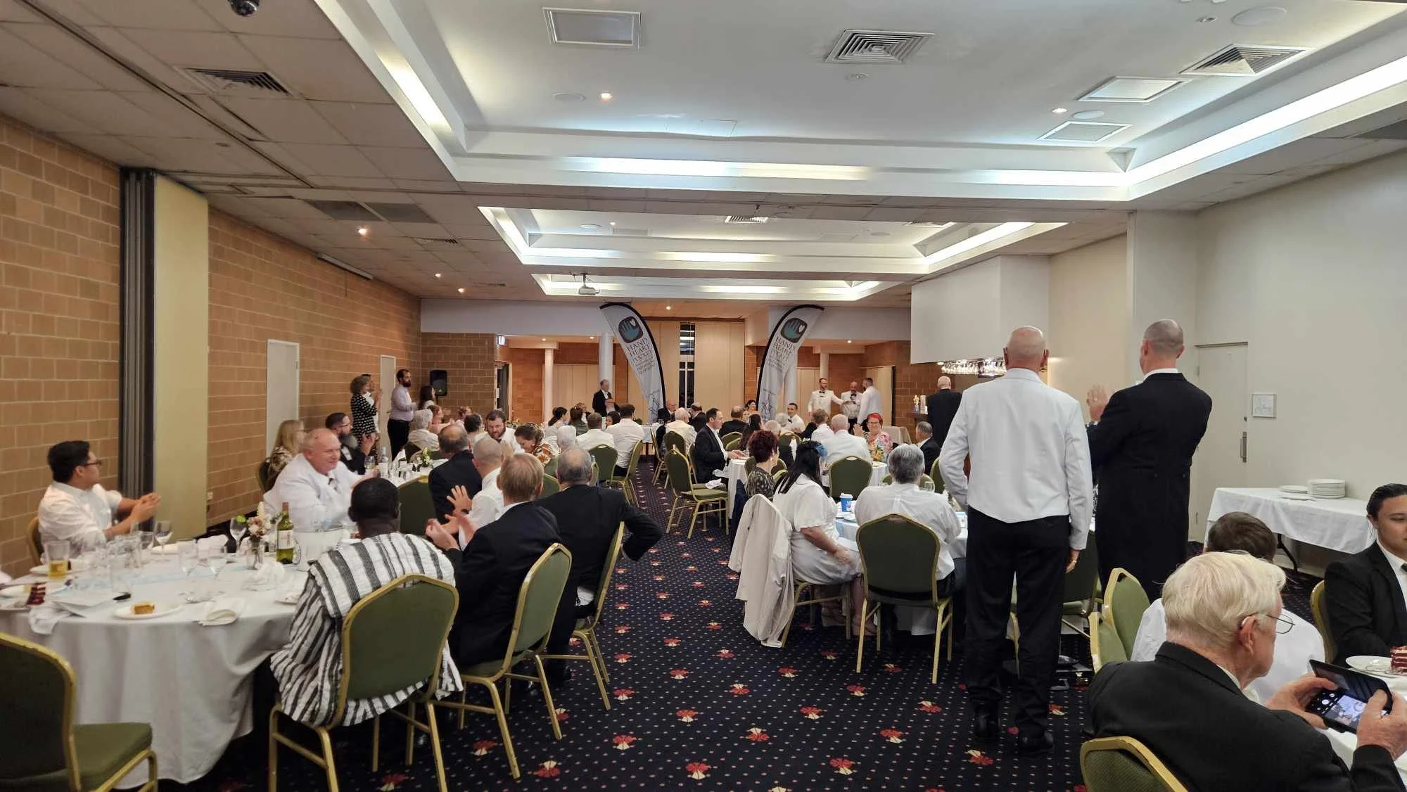 A formal banquet or reception in a hotel conference room with round tables, many guests dressed in formal attire, and a stage at the front with banners and people speaking or preparing to speak.