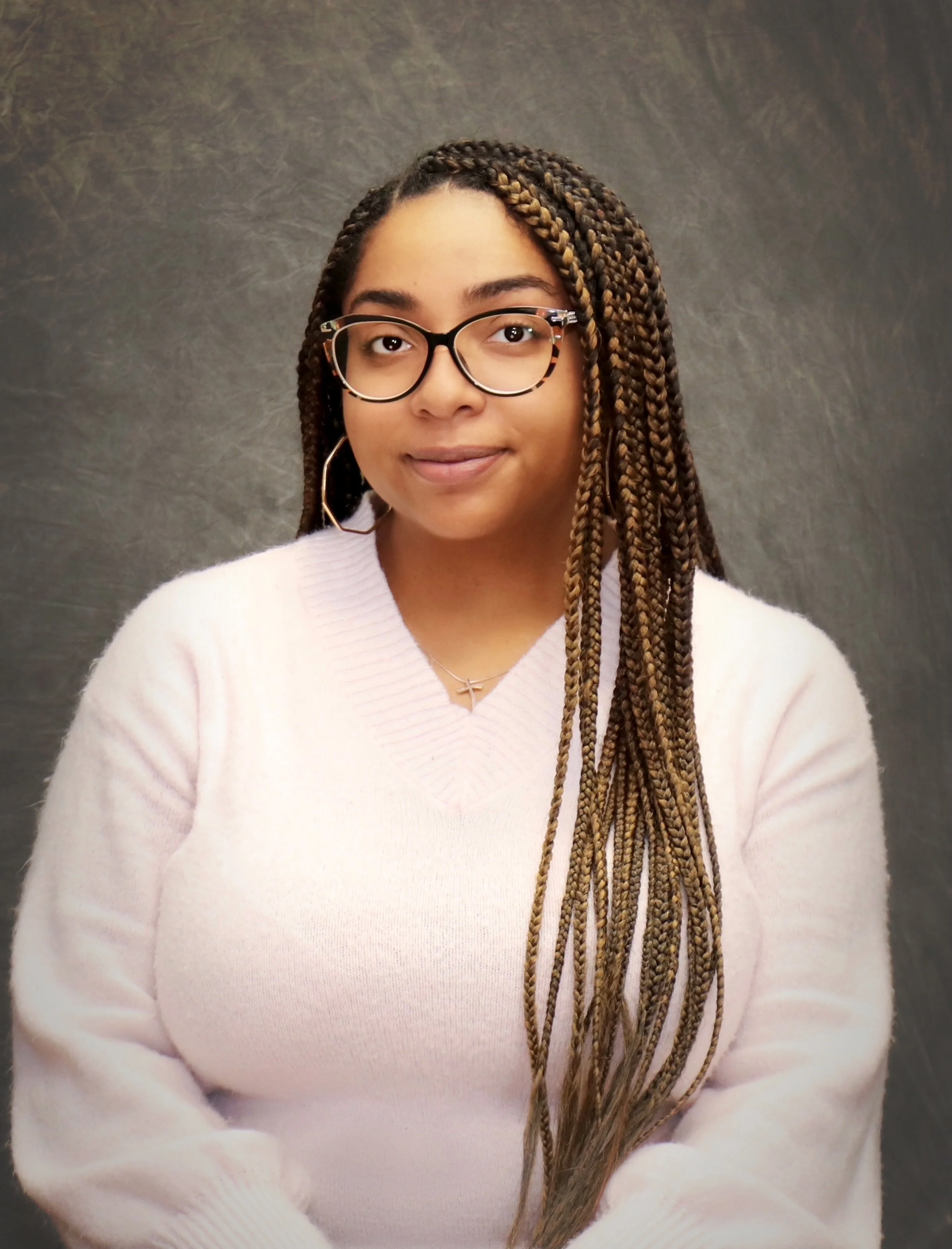 A young woman with long braided hair, glasses, and a light pink sweater, standing against a dark background.