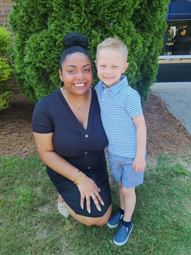 A woman and a young boy standing together outdoors in front of a green bush, smiling at the camera.