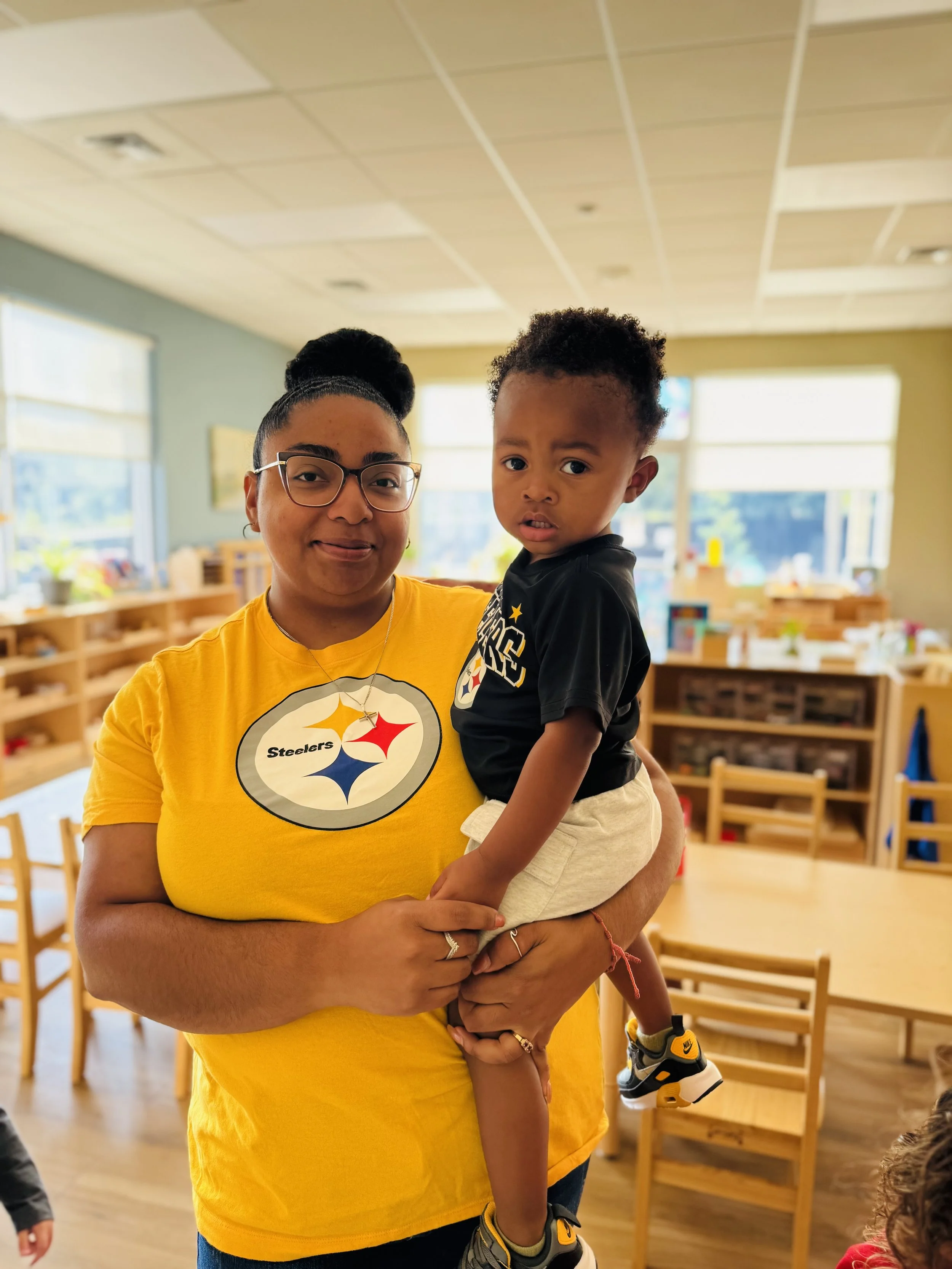 A woman wearing glasses and a yellow Pittsburgh Steelers T-shirt is holding a young child with a concerned expression. The background shows a classroom with wooden chairs and large windows.