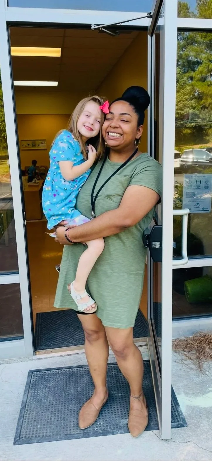 A woman is standing at the entrance of a building, holding a young girl who is smiling and leaning into her. The woman is wearing a green dress, beige shoes, and has her hair in a bun. The girl is wearing a blue dress with a pink bow in her hair. They are both smiling with their eyes closed.