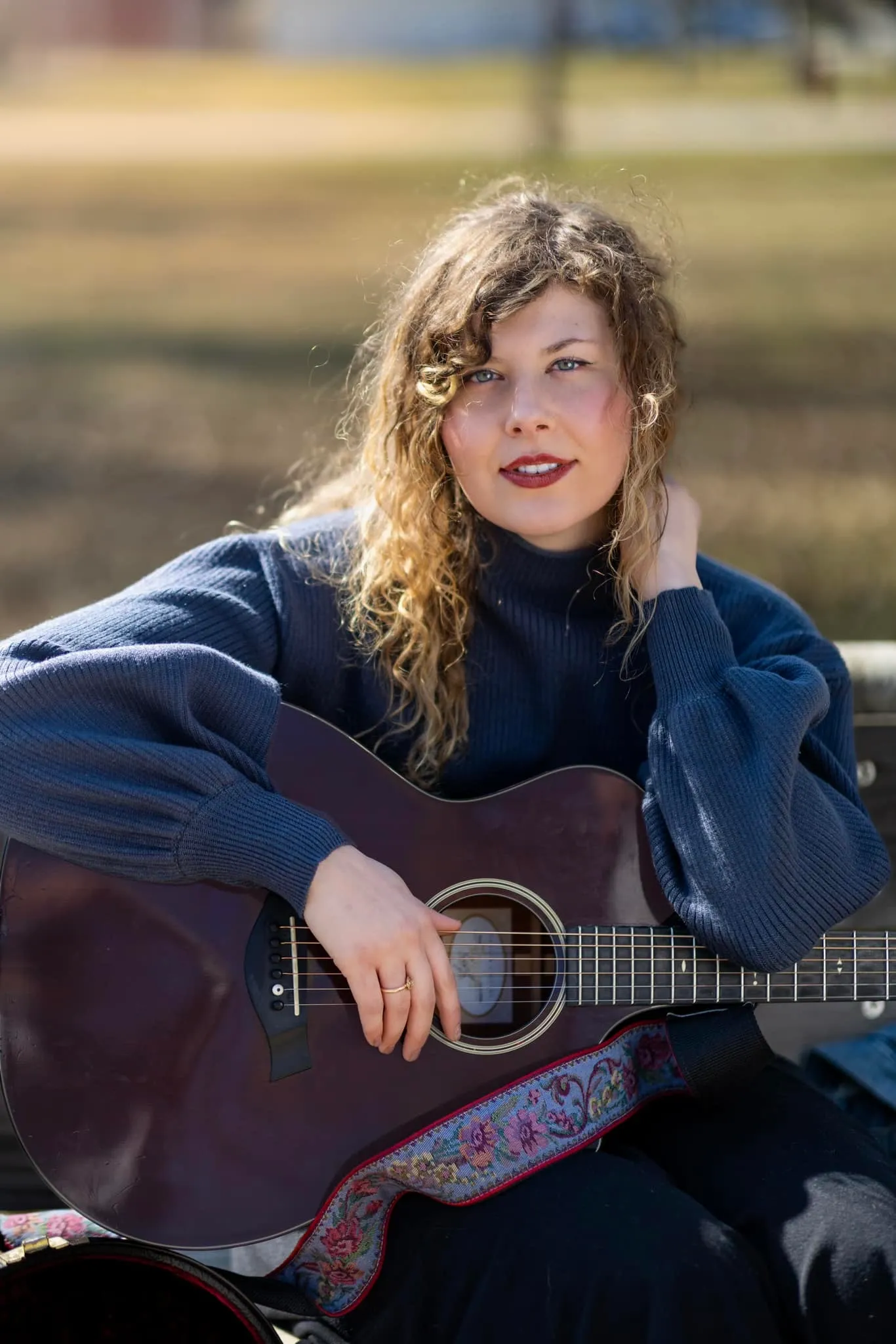 A woman with curly blonde hair and blue eyes sitting outdoors, holding an acoustic guitar with a colorful strap.
