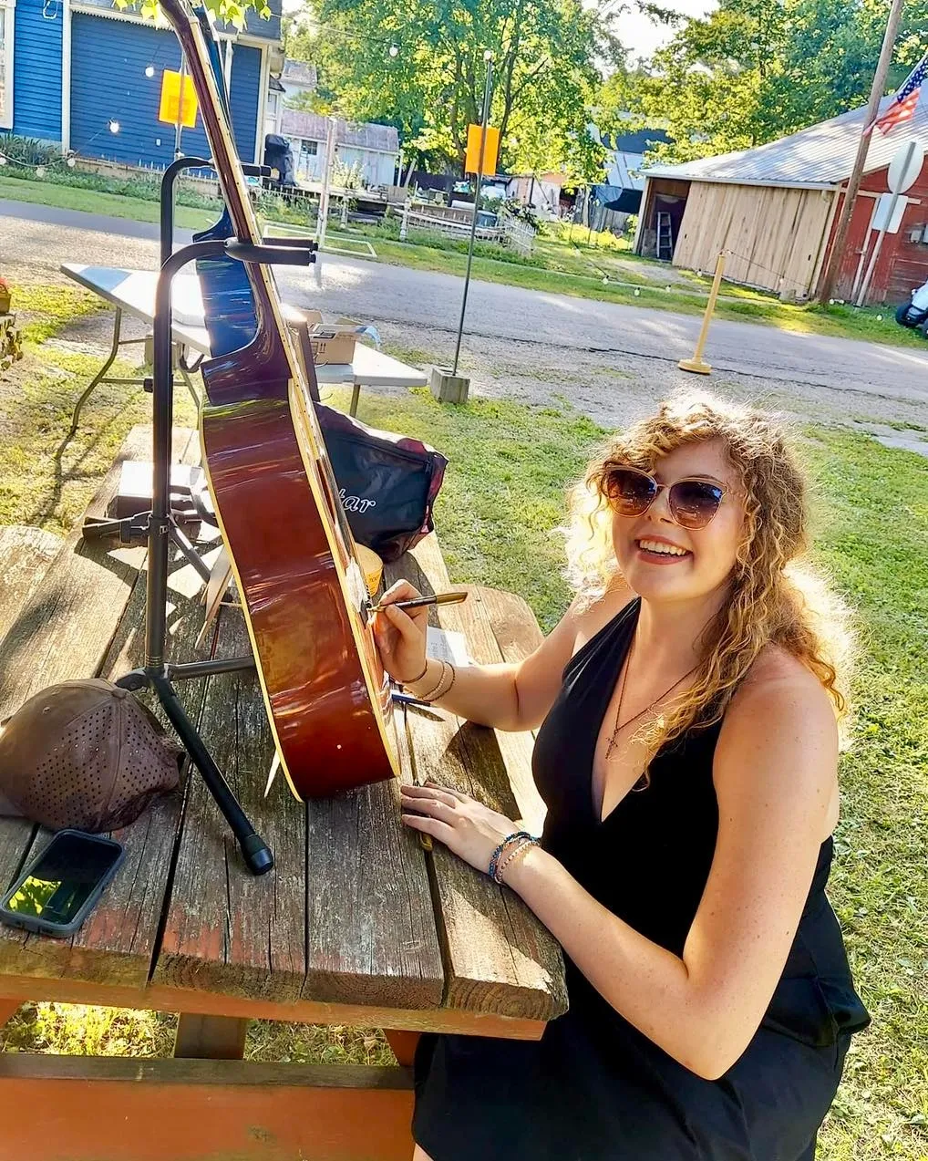 A woman with curly blonde hair, wearing sunglasses and a black dress, is sitting at a wooden picnic table outdoors, signing a guitar with a marker. She is smiling and enjoying a sunny day.