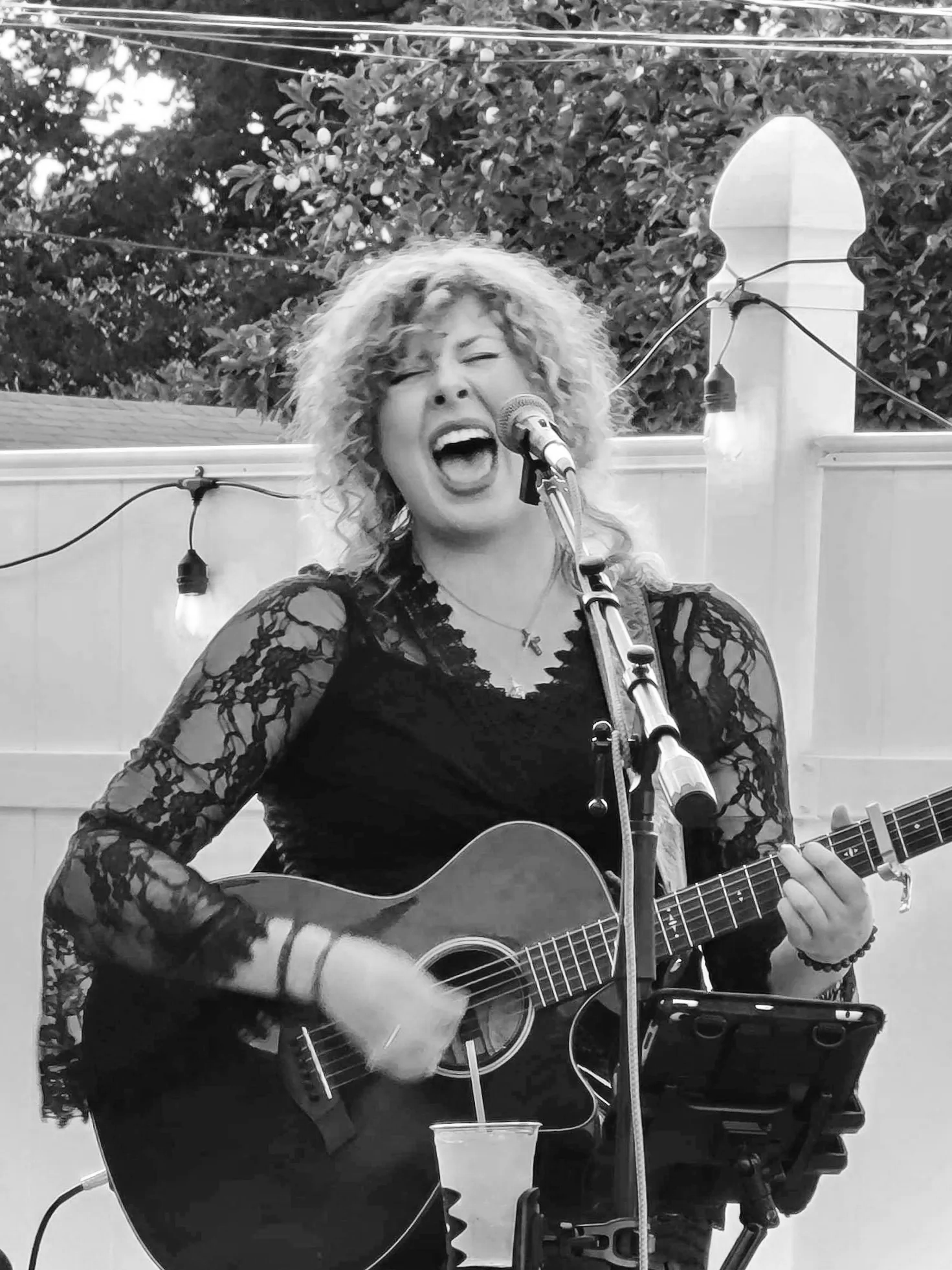 A woman with curly hair singing passionately into a microphone while playing an acoustic guitar on an outdoor stage, with string lights and trees in the background.