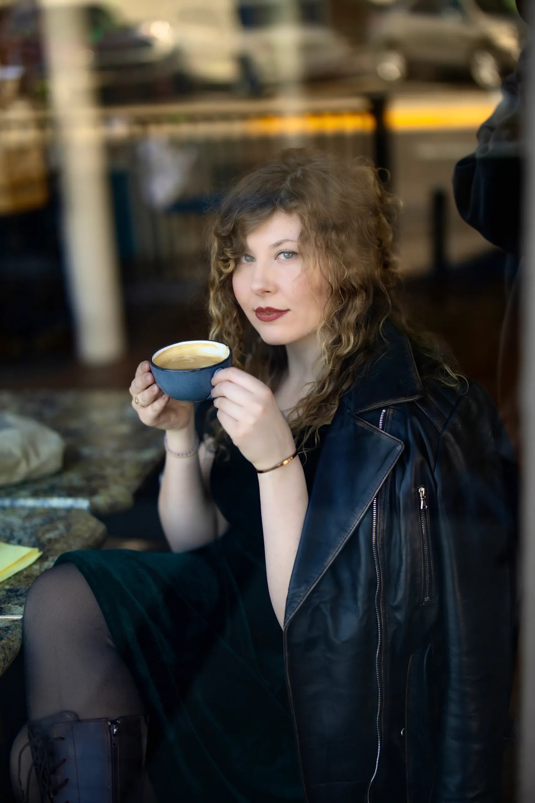 A young woman with curly hair and fair skin sitting at a cafe, holding a cup of coffee and looking at the camera, wearing a black leather jacket, dark dress, and black tights.