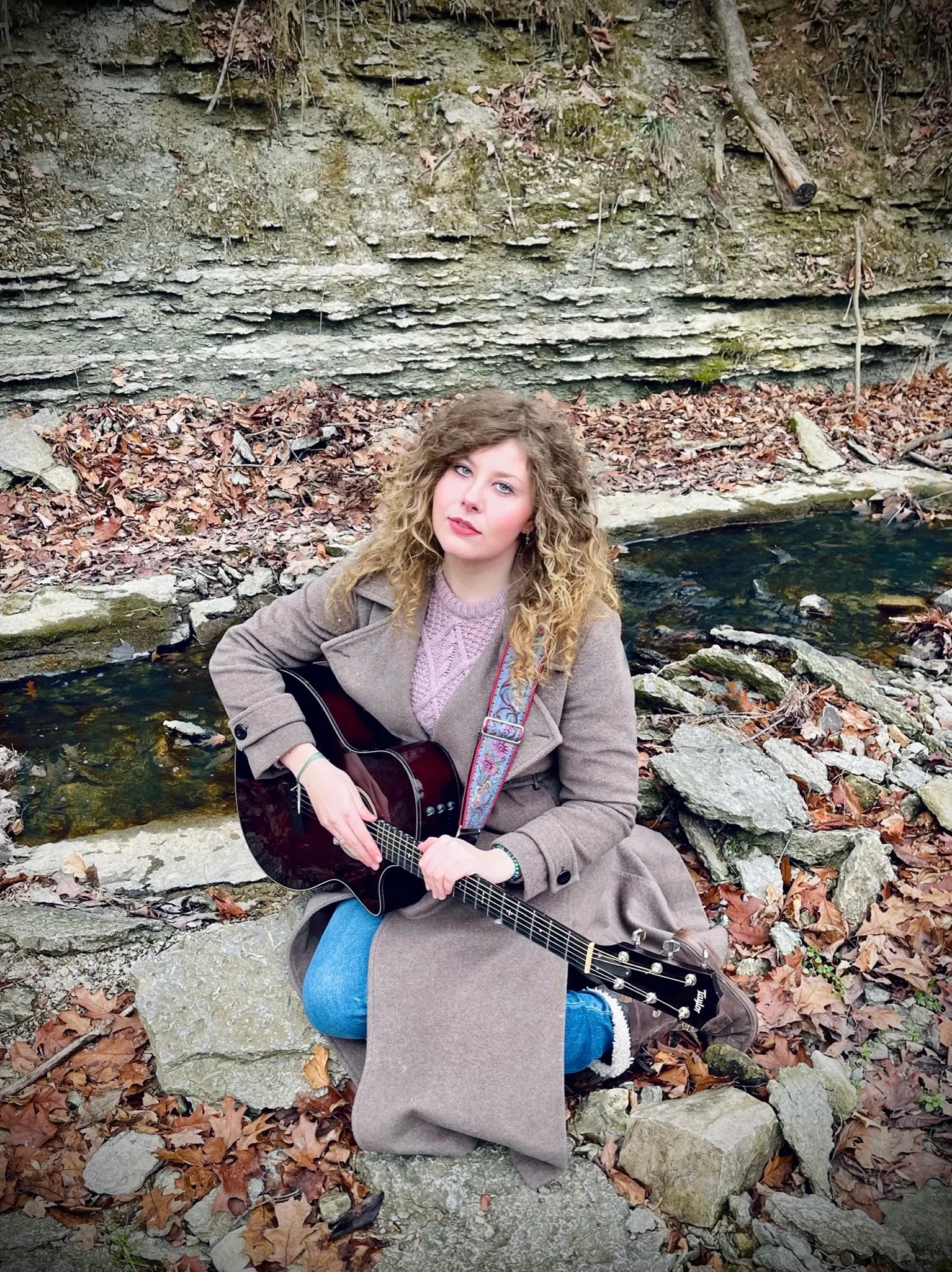 A woman with curly hair sitting on rocks beside a small stream, holding a black acoustic guitar, surrounded by fallen leaves and a rocky background.