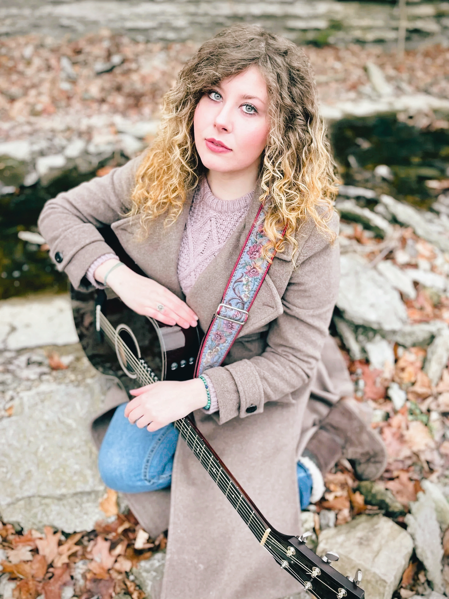 A young woman with curly blonde hair and blue eyes kneels outdoors among rocks and fallen leaves, holding a black acoustic guitar with a colorful strap.