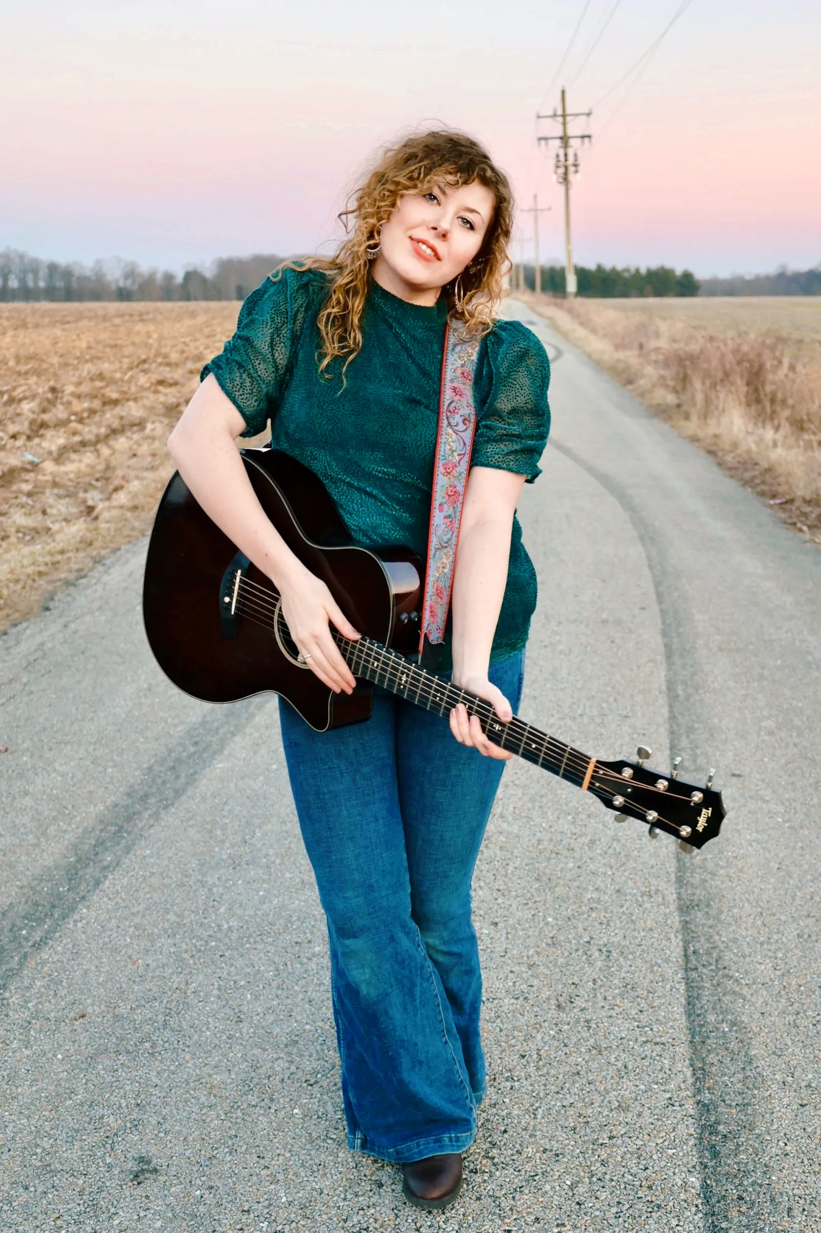Young woman with curly hair playing an acoustic guitar on a rural road at sunset.