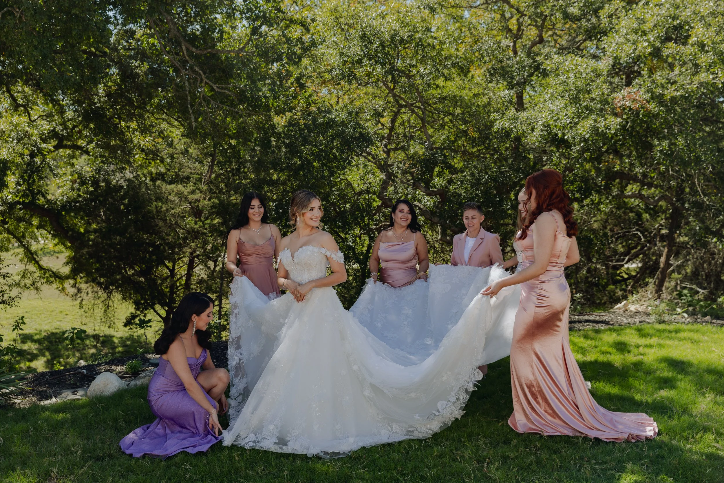 A bride with blonde hair in a white wedding gown surrounded by six bridesmaids in pastel dresses, standing outdoors on a grassy area with trees in the background.