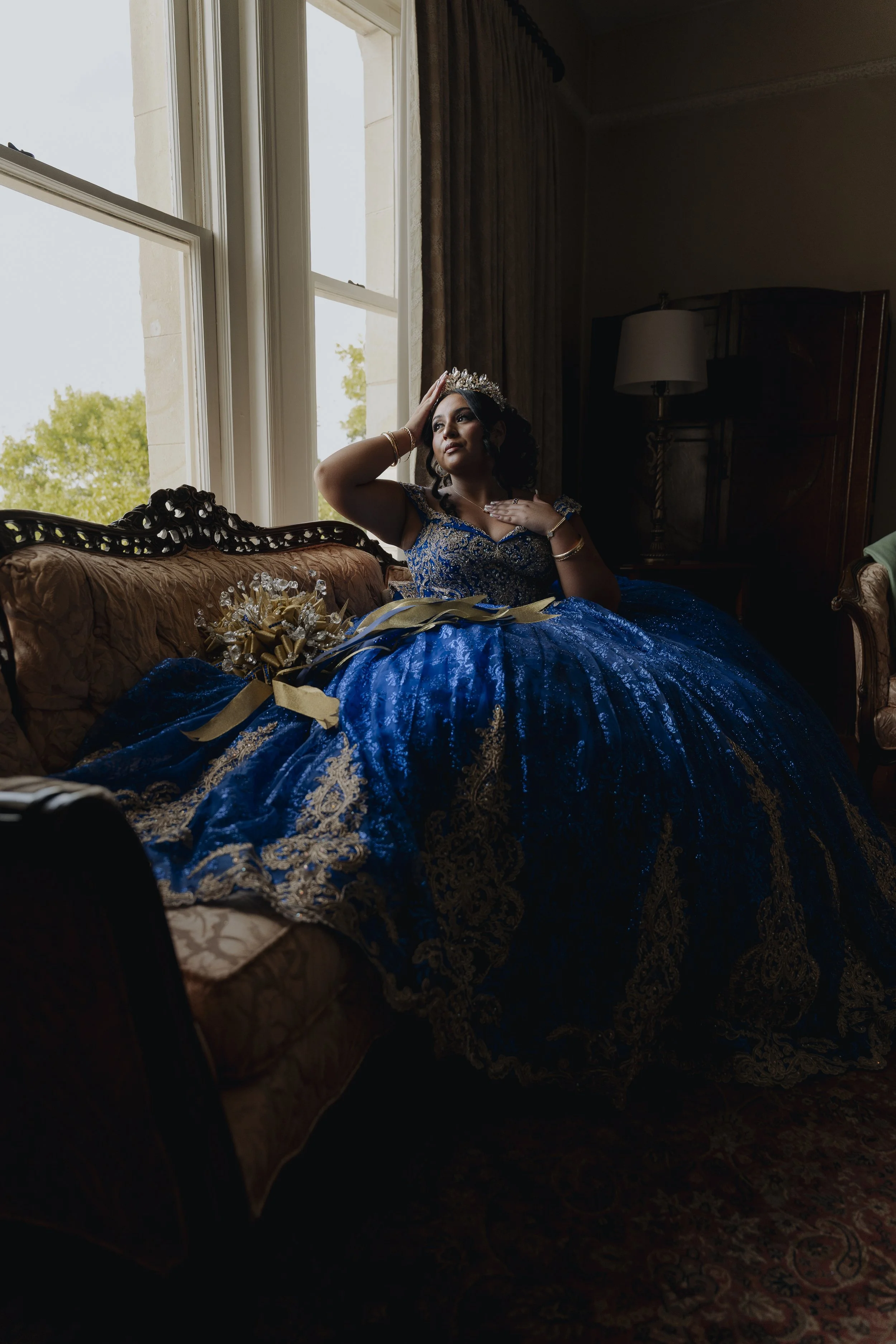 A woman dressed in a luxurious blue and gold gown, sitting on a vintage sofa by a large window, wearing a tiara, with her hand on her forehead, and holding a bouquet of flowers.