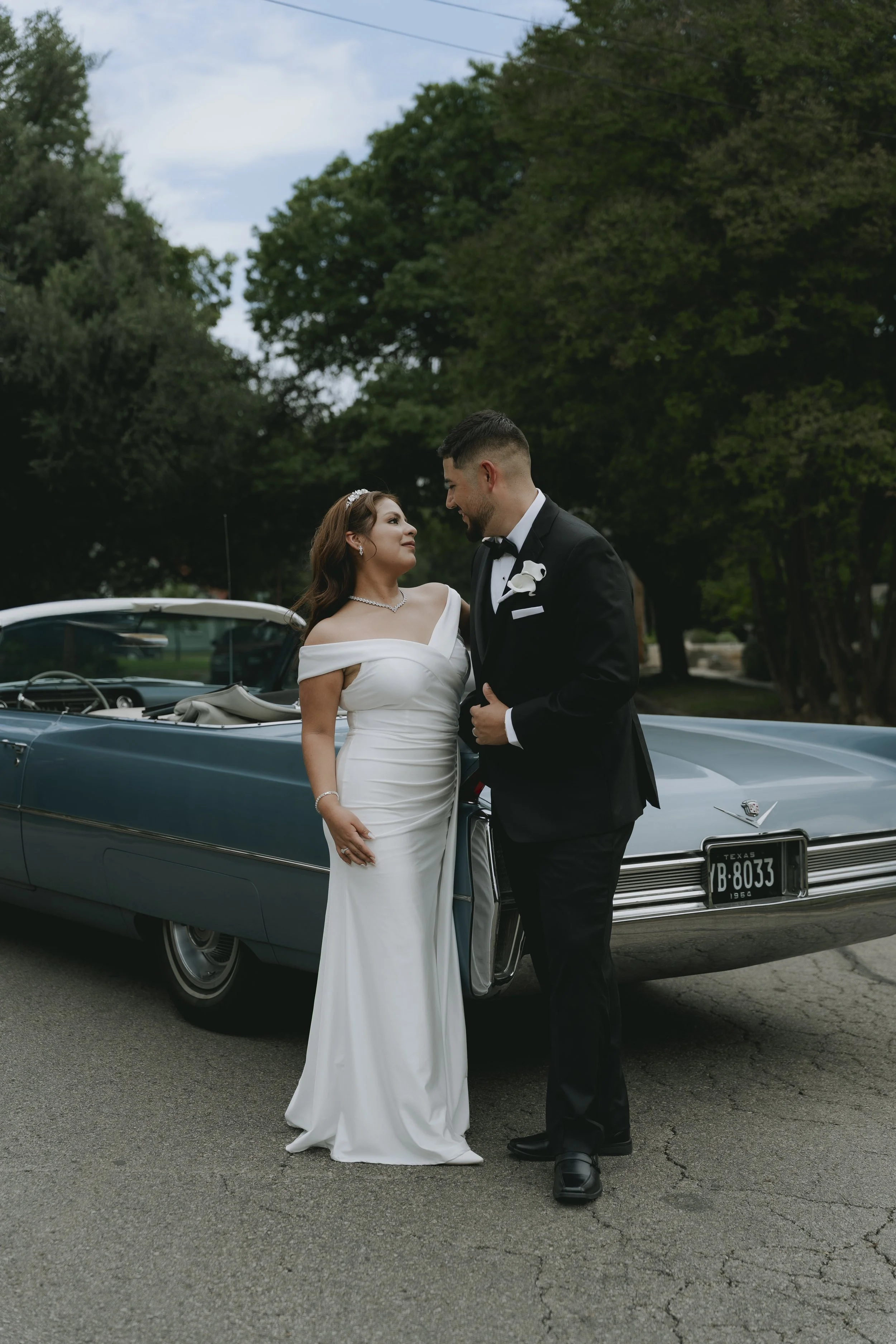 A bride and groom in wedding attire standing next to a vintage blue Cadillac with Texas license plates. The couple is looking at each other affectionately outdoors with green trees and a cloudy sky in the background.