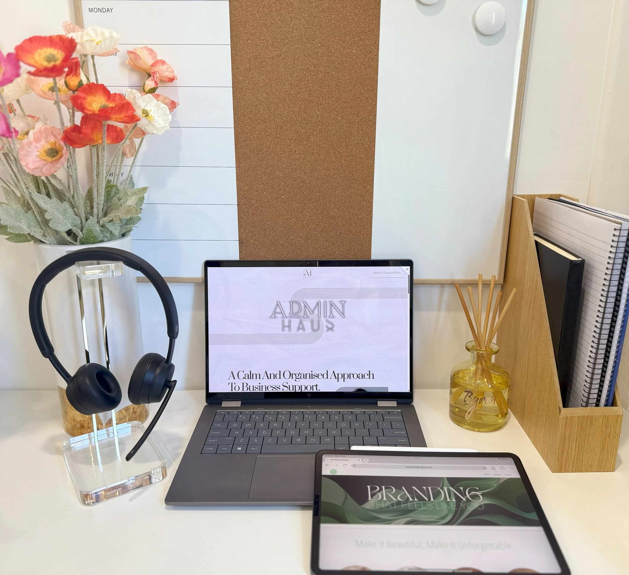 Office desk with a laptop displaying 'ADMIN HAUS' and the tagline 'A Calm And Organised Approach To Business Support,' a tablet showing 'BRANDING' and the phrase 'Make It Beautiful, Make It Unforgettable,' a vase with reed diffusers, a pair of black headphones on a stand, a whiteboard with a cork section, and a floral arrangement of pink, white, and red flowers.