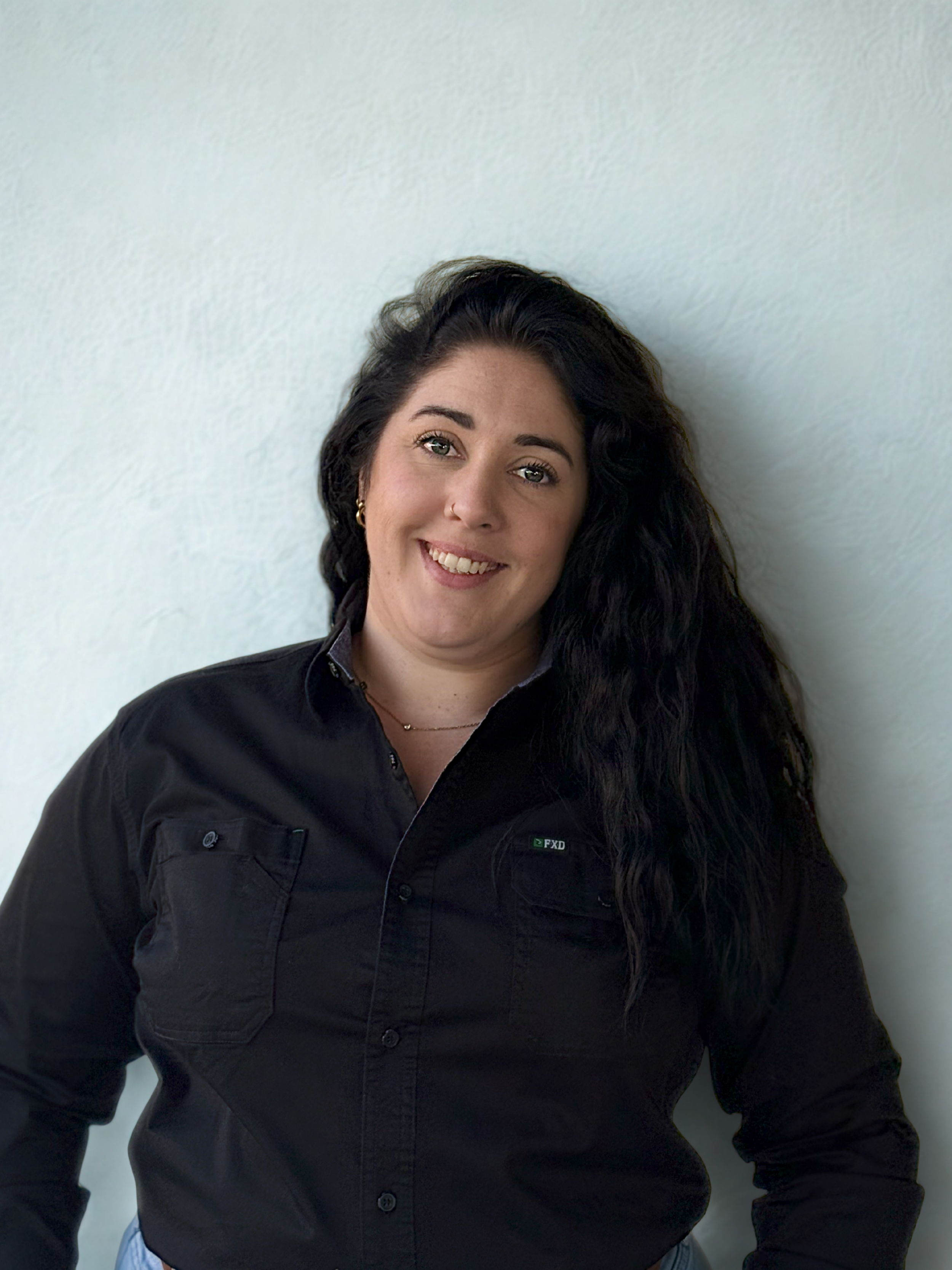 A woman with long, dark, curly hair smiling in front of a plain, light-colored wall.