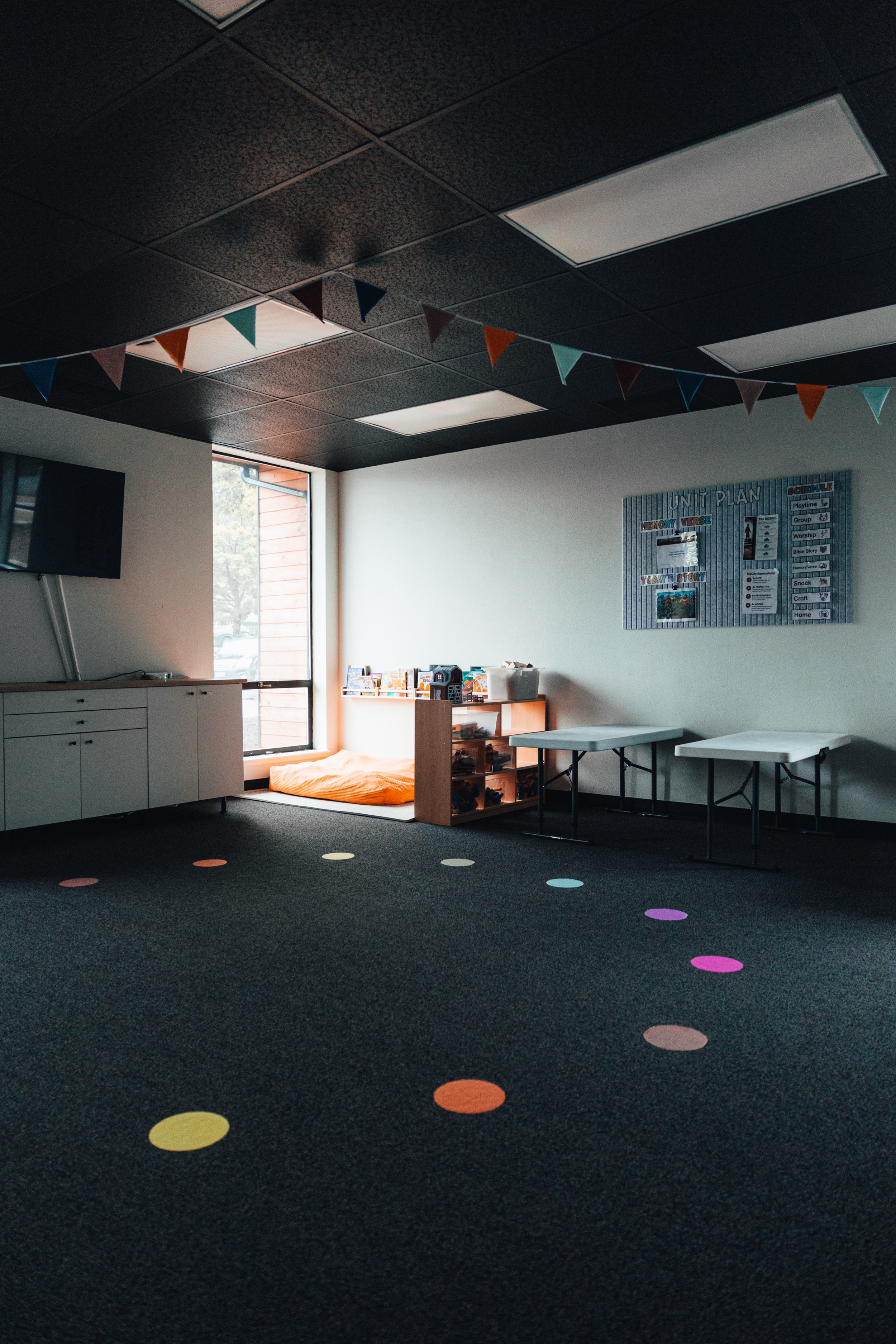 Empty classroom with black carpet, colorful dots, bunting on ceiling, and a large window allowing natural light, with tables, shelving, and a bulletin board.