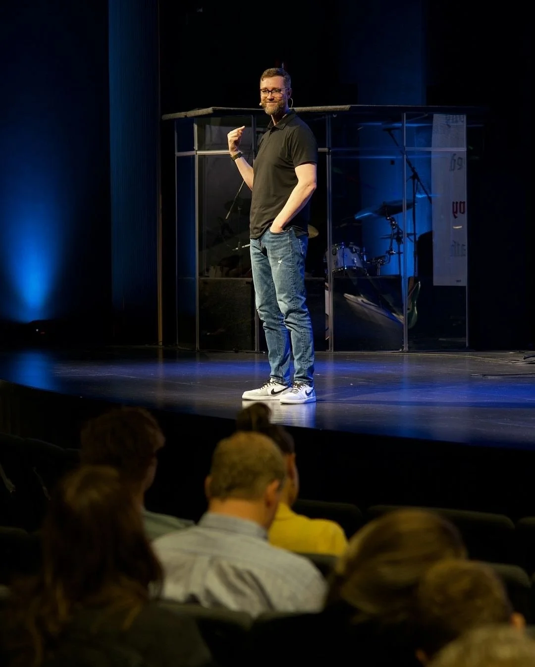 A man in a black polo shirt, jeans, and white Nike sneakers giving a presentation on stage in front of an audience, with a drum set and microphone in the background.