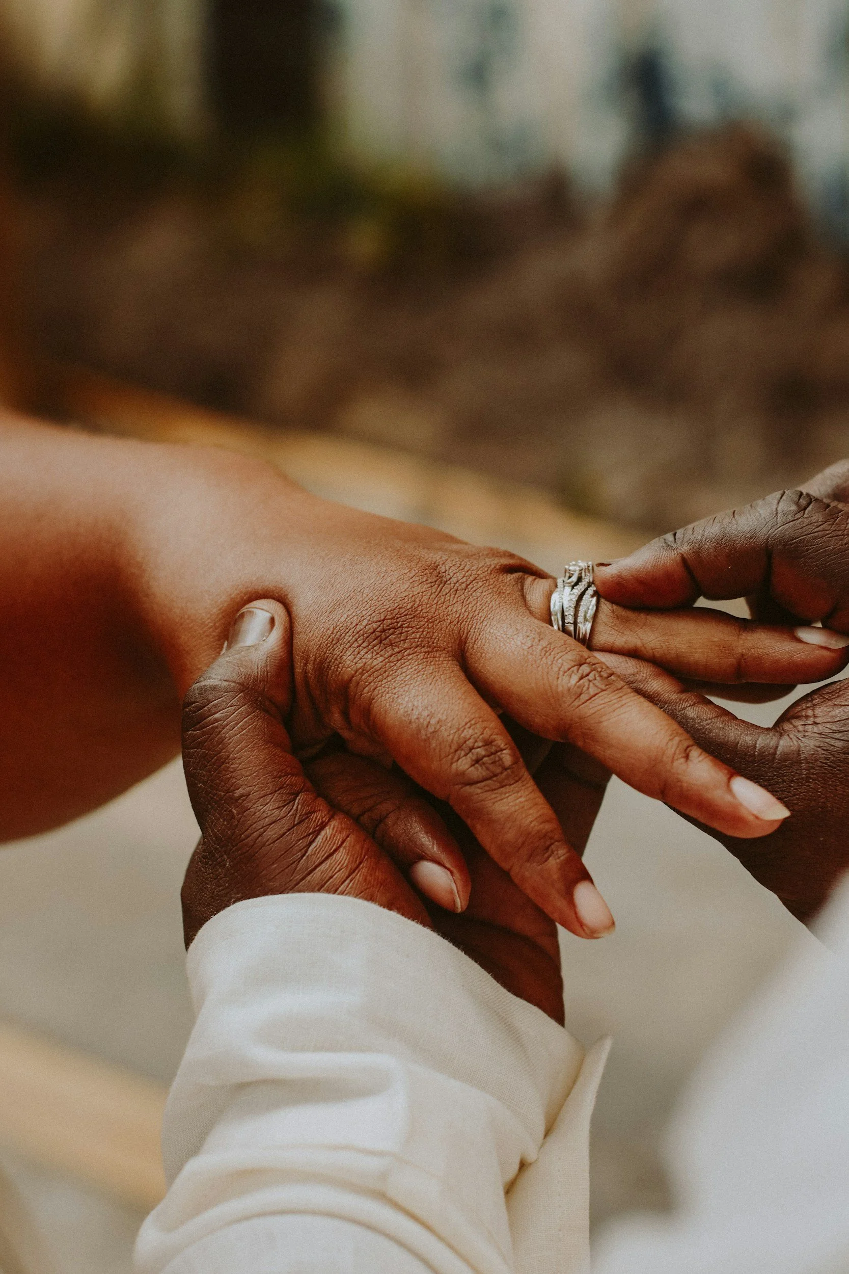 Two hands, one dark-skinned and one light-skinned, holding each other, with the light-skinned person placing a ring on the dark-skinned person's finger. The scene suggests a moment of engagement or wedding.