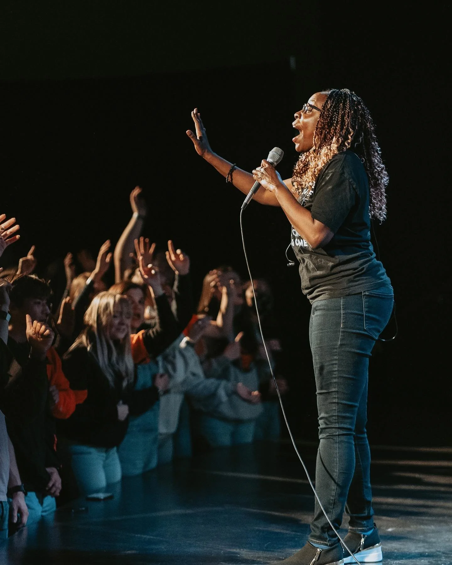 A woman with braids and glasses holding a microphone, speaking or singing to a lively audience with hands raised.