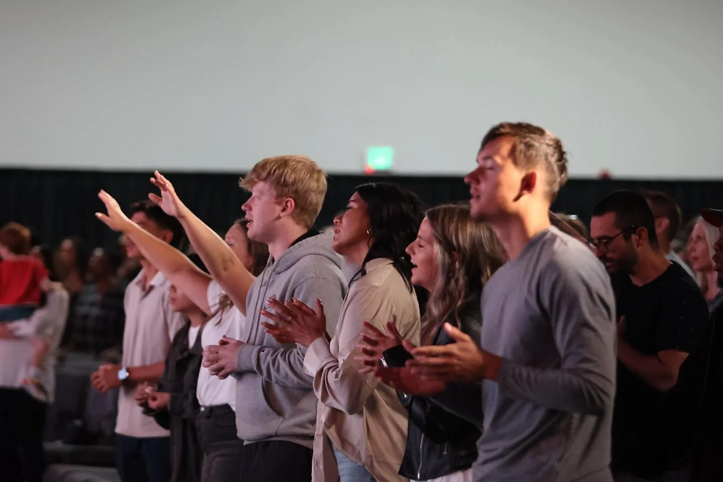 People standing in a congregation, some with hands raised and others clapping, during a lively event or service in a large indoor venue.